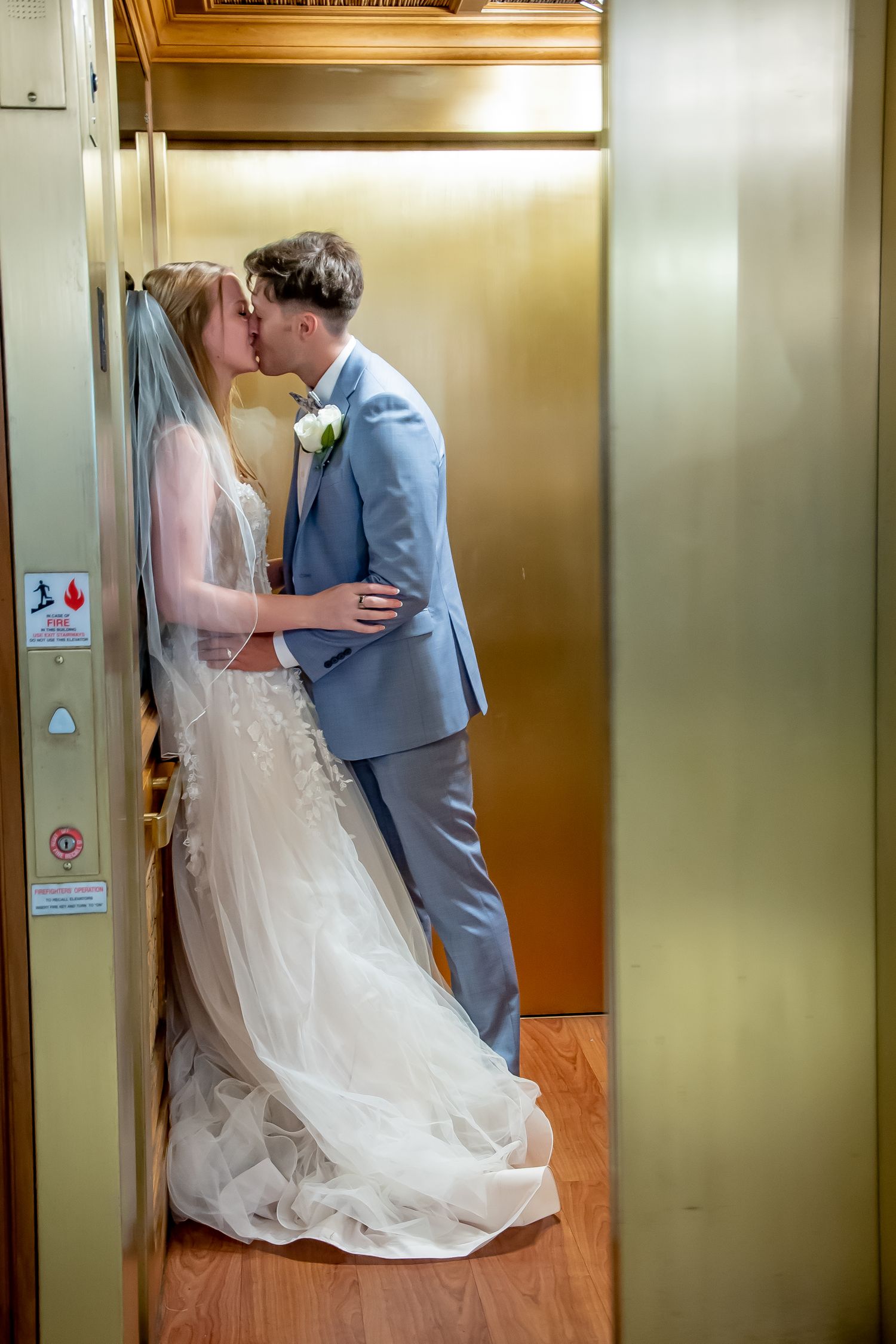 Bride and groom sharing a romantic kiss inside a golden elevator, bride wearing white gown with veil, groom in light blue suit.