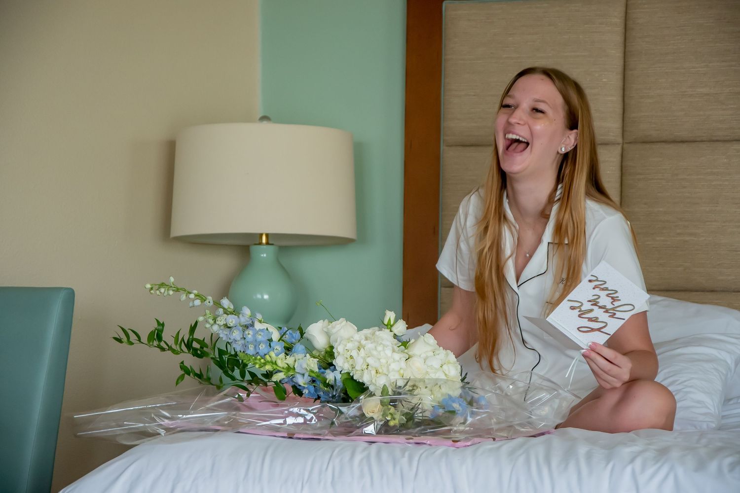 Laughing bride in white pajamas sits on hotel bed holding card with blue and white flower bouquet.