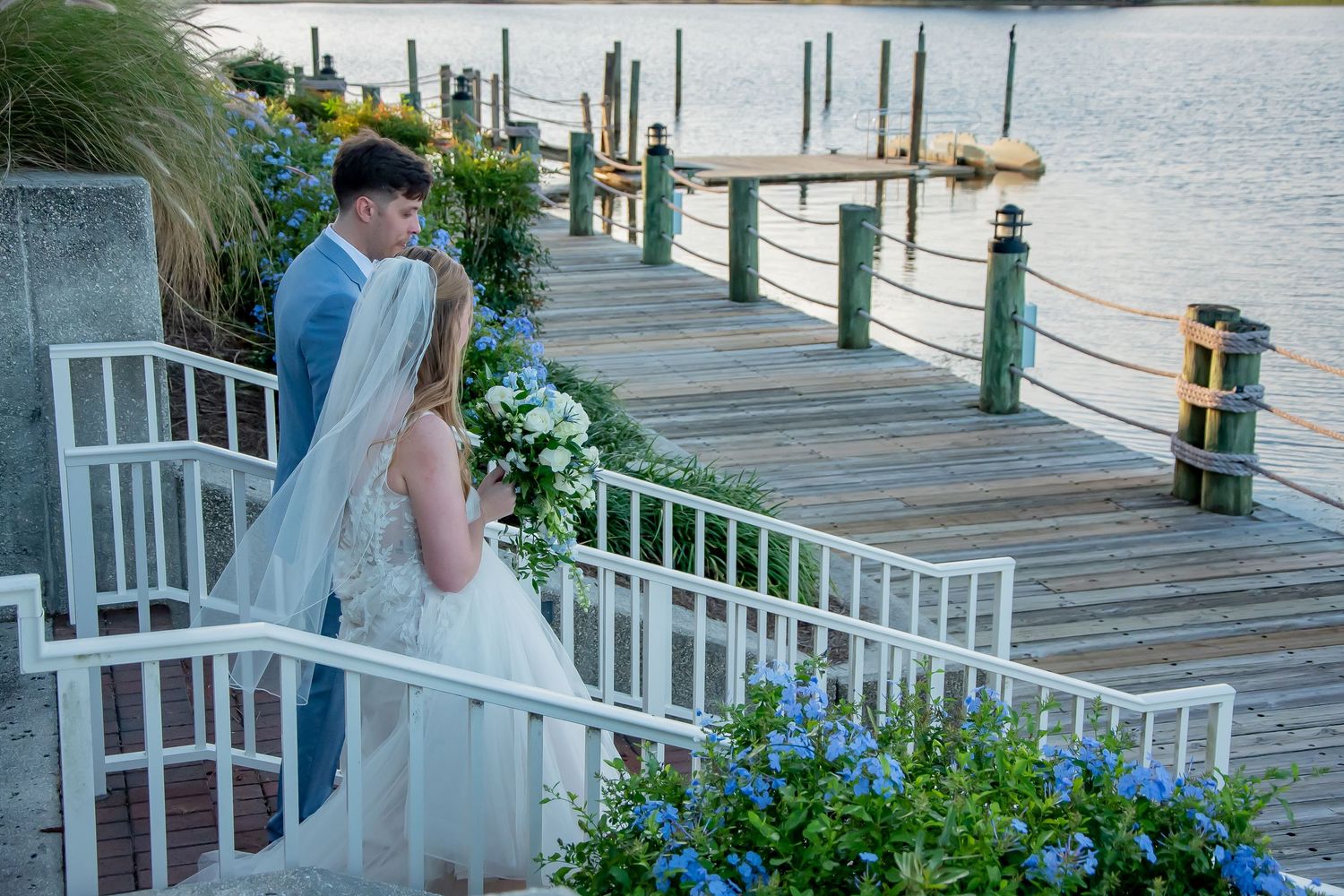 Bride and groom share a romantic moment on a waterfront dock adorned with white flowers and lush greenery.