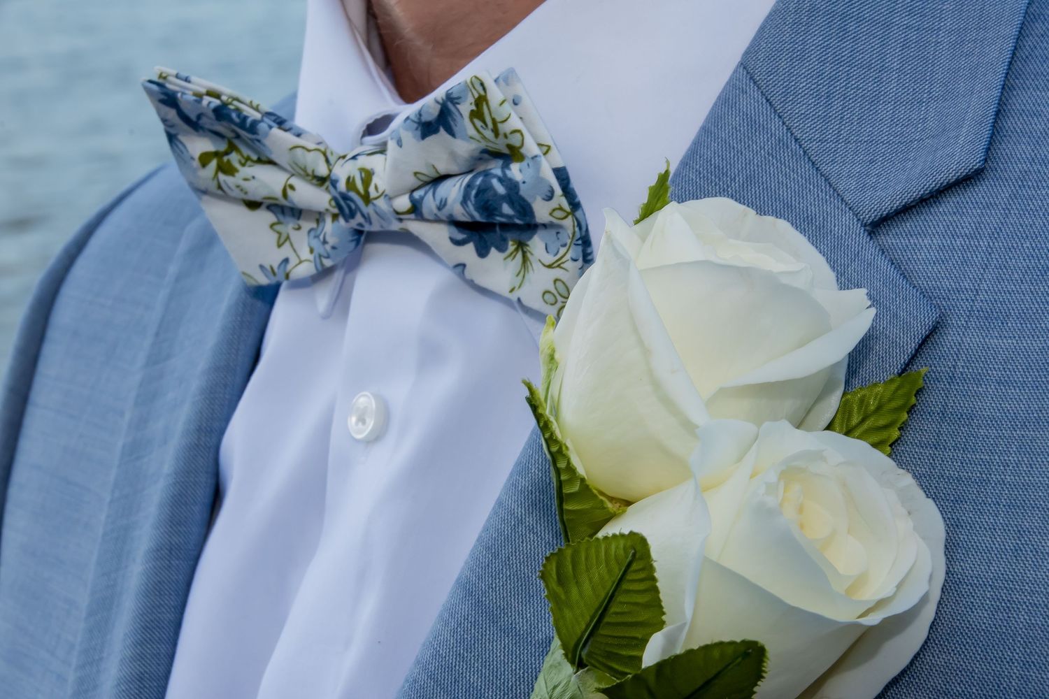 Close-up of a groom wearing a floral blue bow tie and white rose boutonniere on a light blue suit jacket.