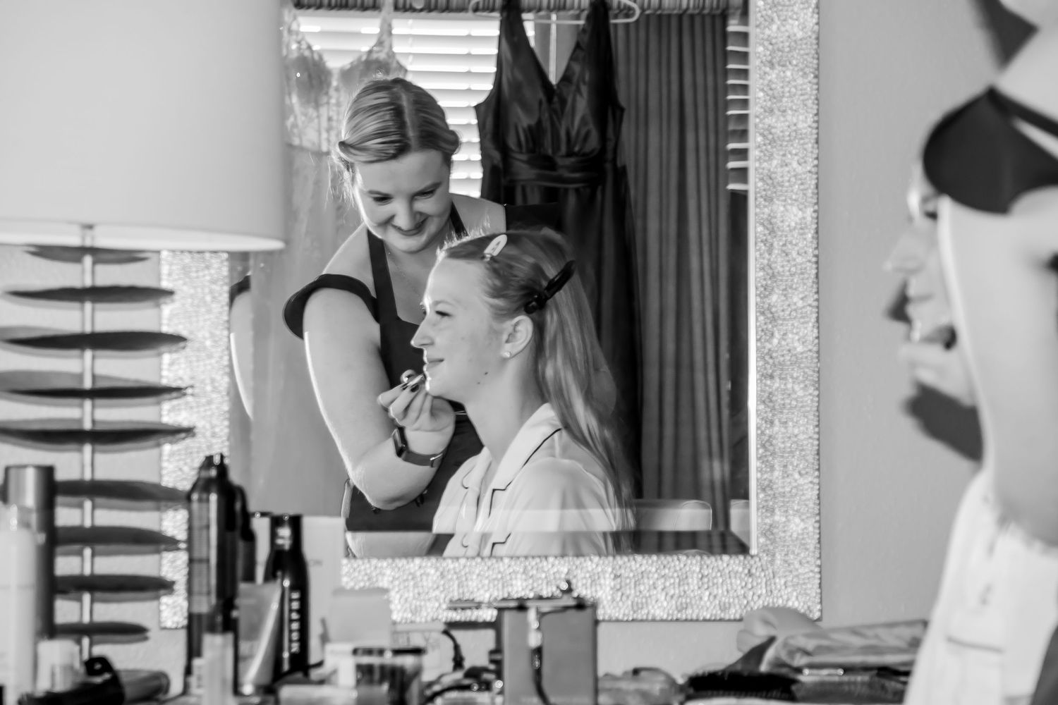 Black and white photo of a makeup artist applying makeup to a bride while smiling in a mirror reflection.