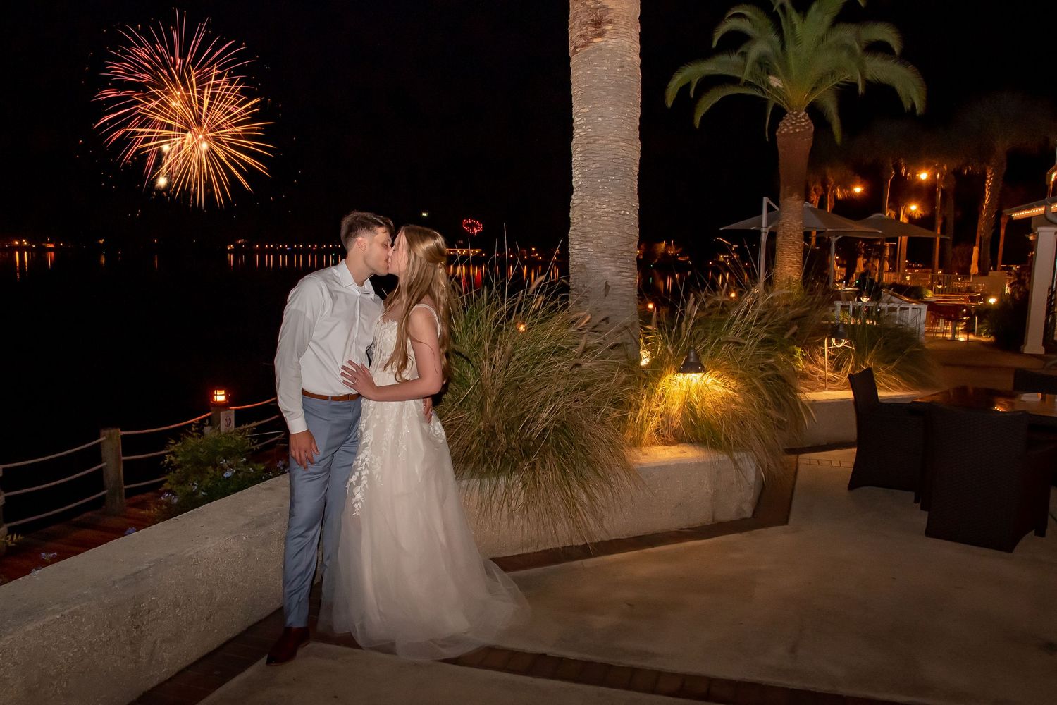 Bride and groom share a romantic kiss on a waterfront terrace with colorful fireworks lighting the night sky.