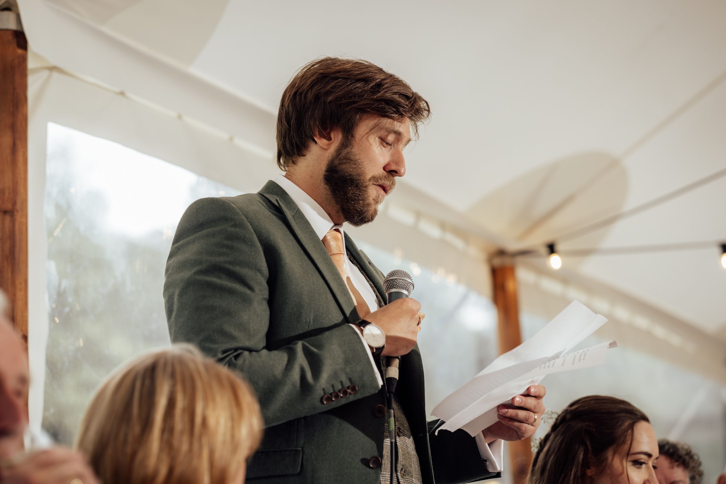 A bearded man in a green suit delivers a speech at a wedding reception, holding a microphone and notes.