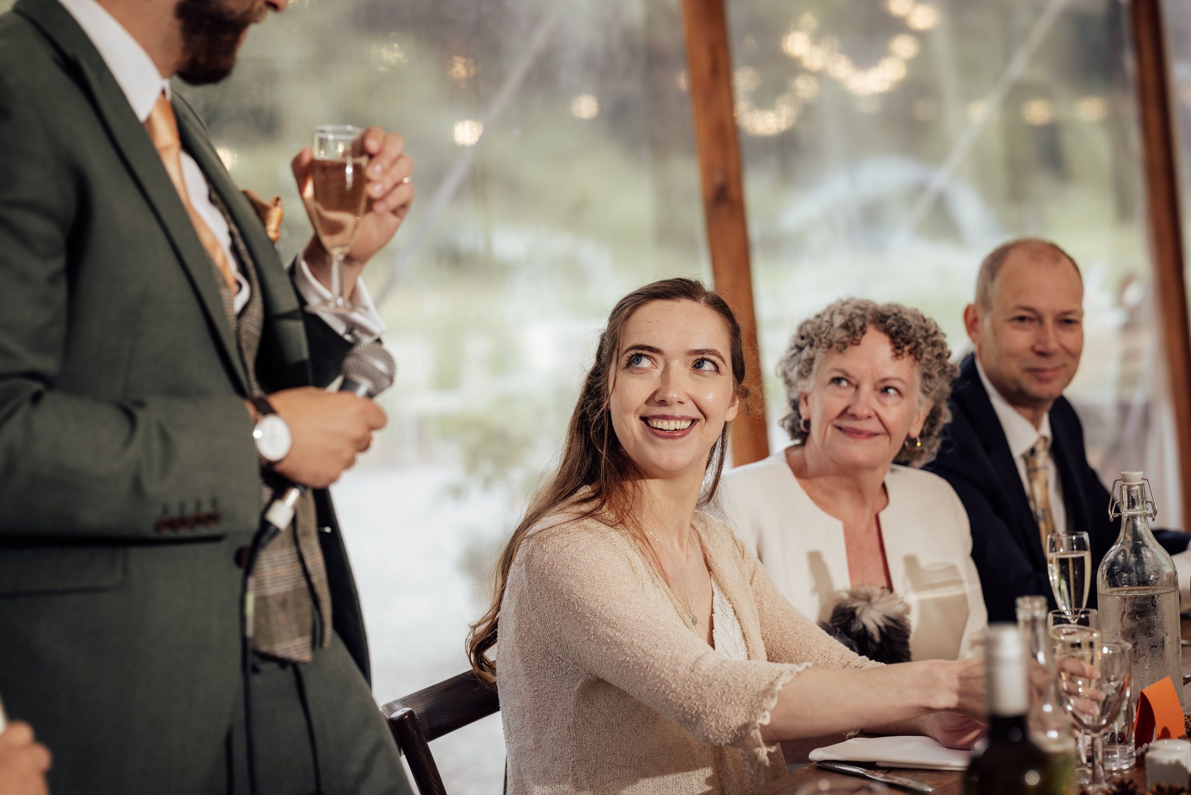 Bride smiling joyfully at wedding reception table with guests holding champagne glasses in elegant venue.