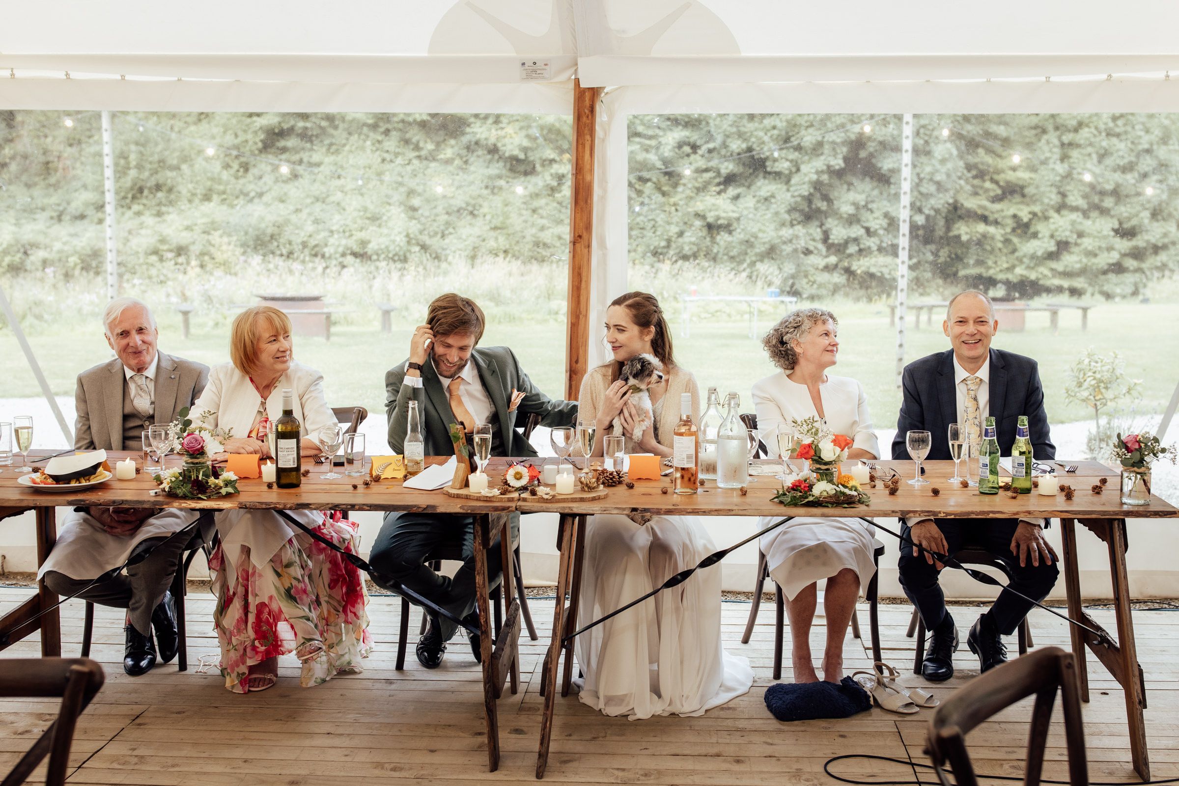 Wedding party seated at a long rustic table under a marquee tent, surrounded by floral arrangements and drinks.
