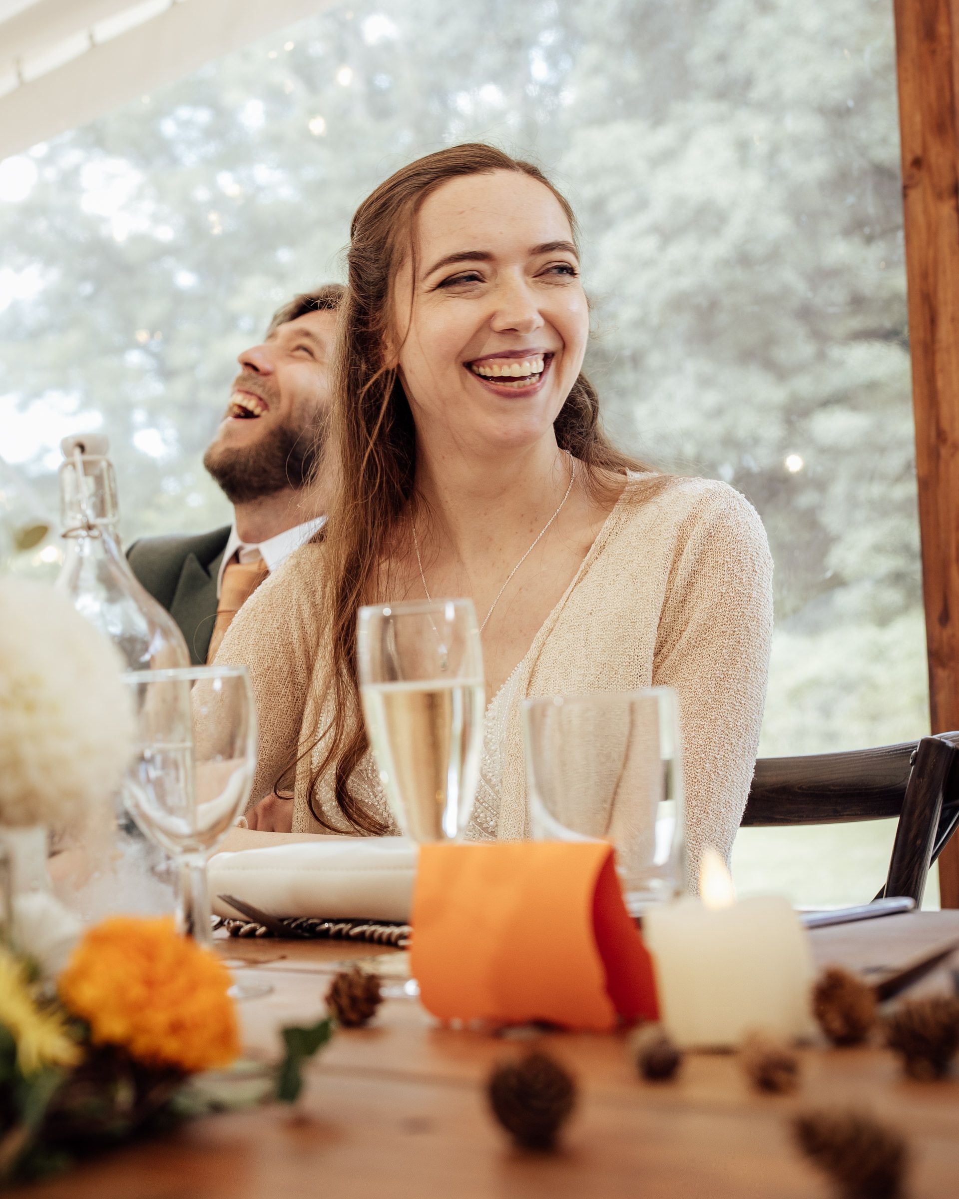 A joyful woman laughs at a elegantly set wedding reception table with champagne glasses and floral centerpieces.