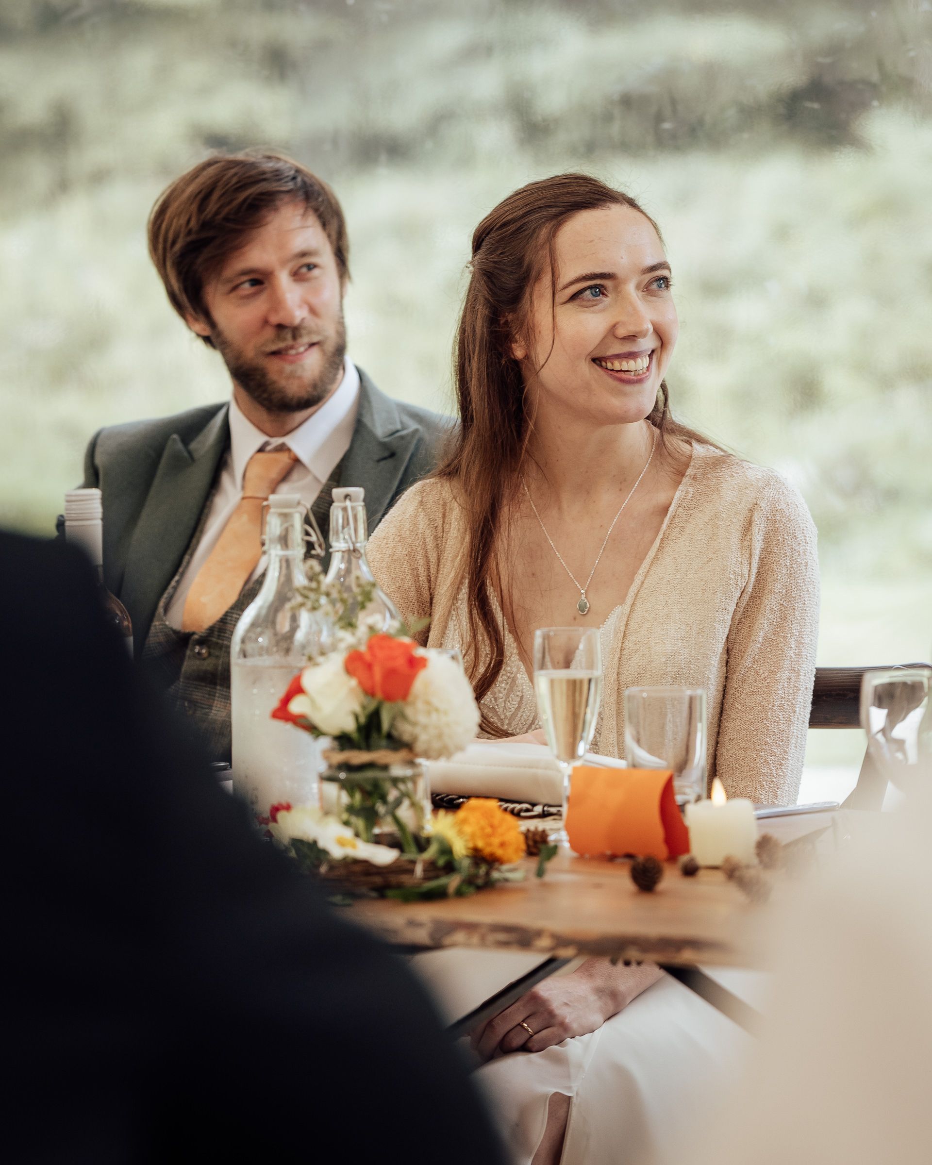 A smiling couple sits at a decorated wedding reception table with floral centerpieces and candles.