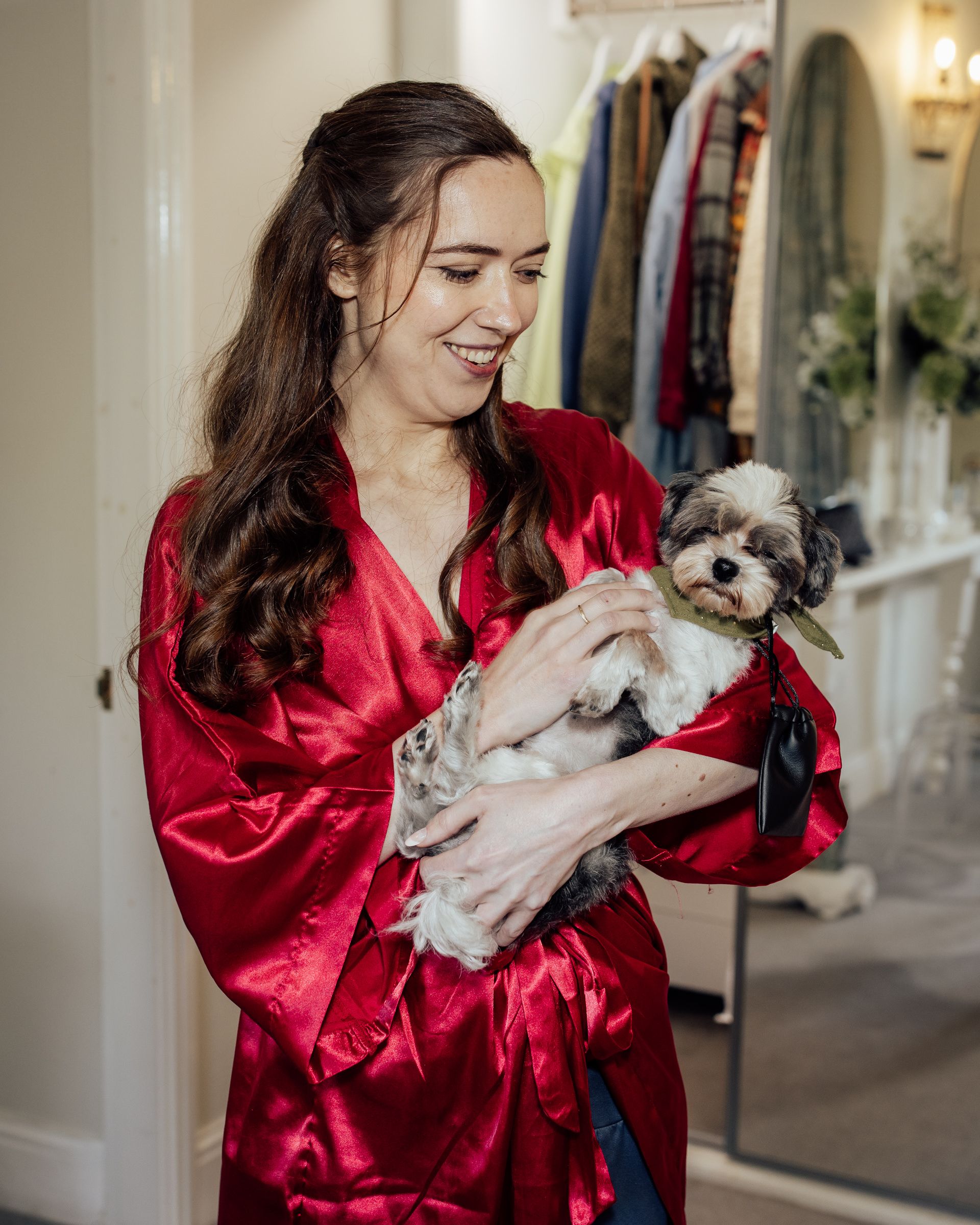 Woman in red satin robe smiling while holding a small Shih Tzu dog indoors near a clothing rack.