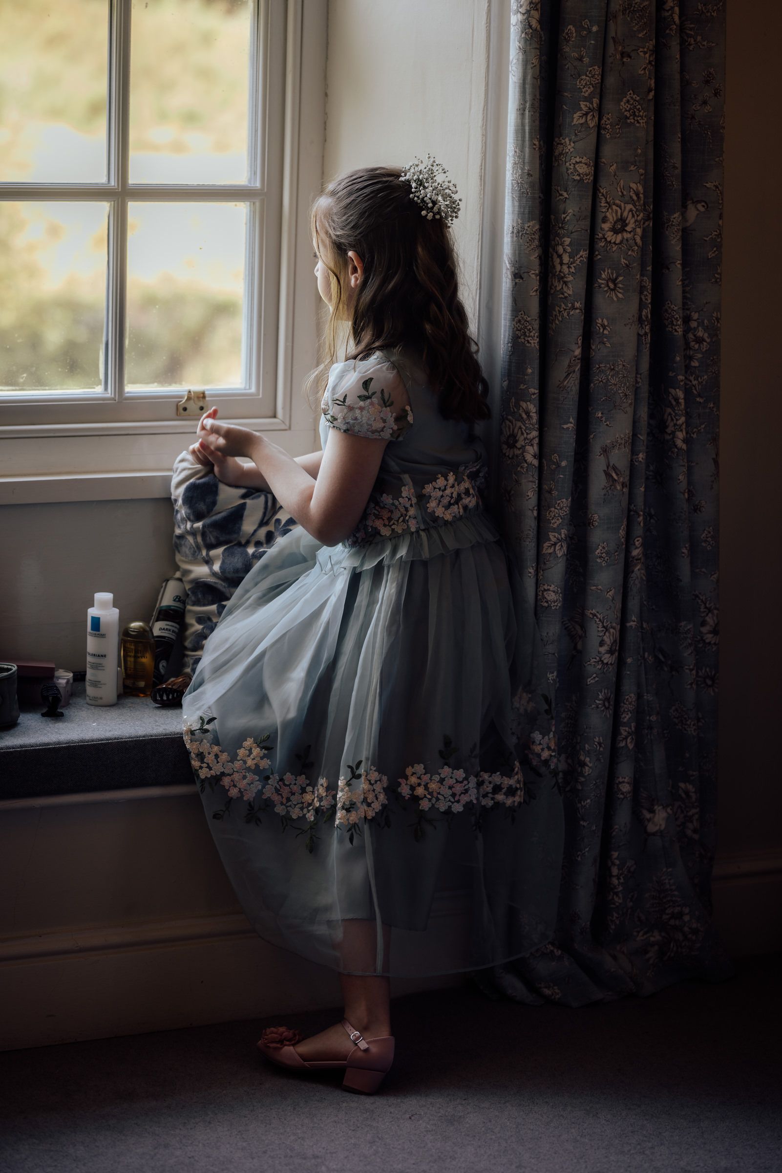 Young girl in floral tulle dress with hair accessory gazes out a window beside elegant curtains.