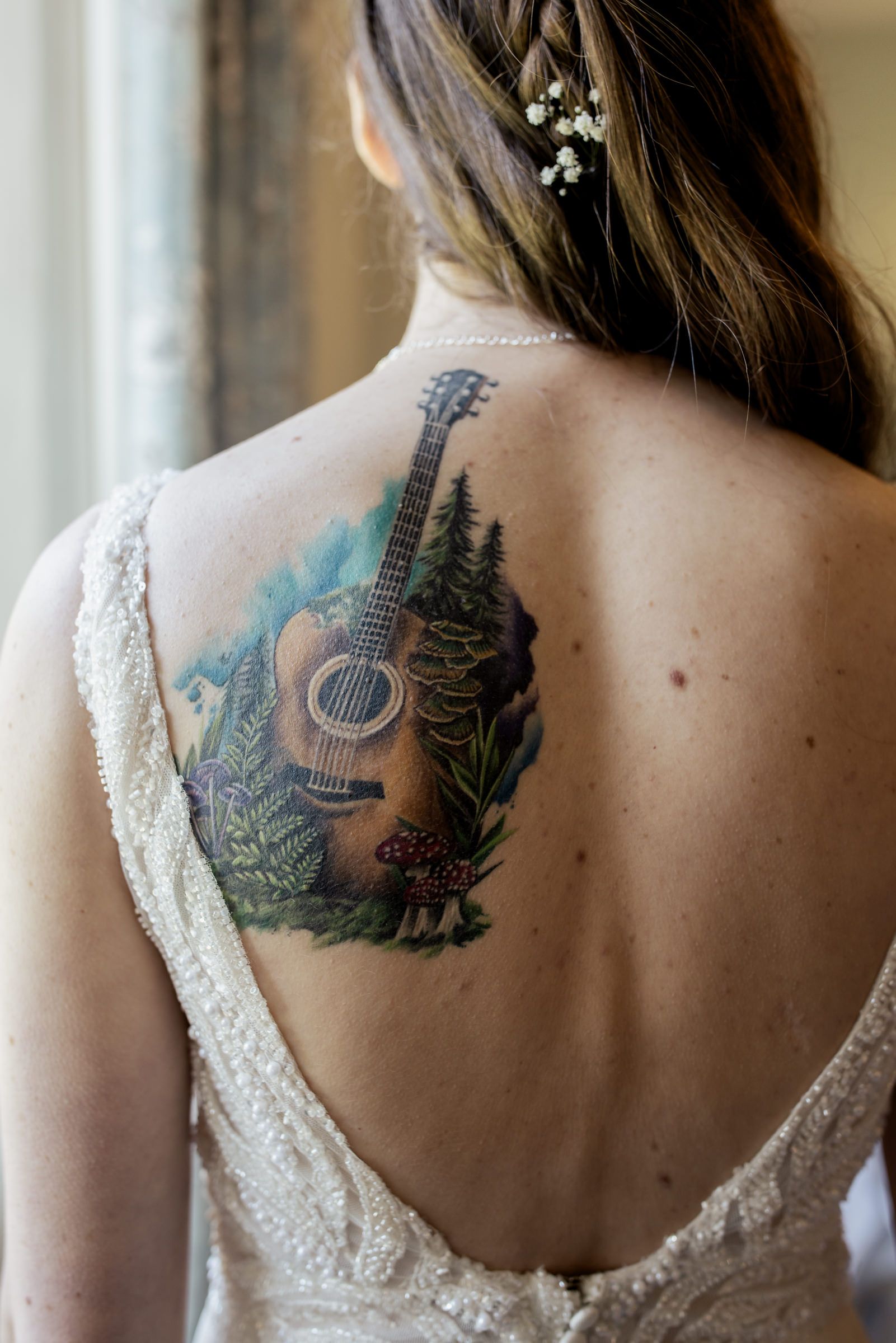 Bride in lace wedding dress showing back tattoo of acoustic guitar with forest mushrooms and greenery.