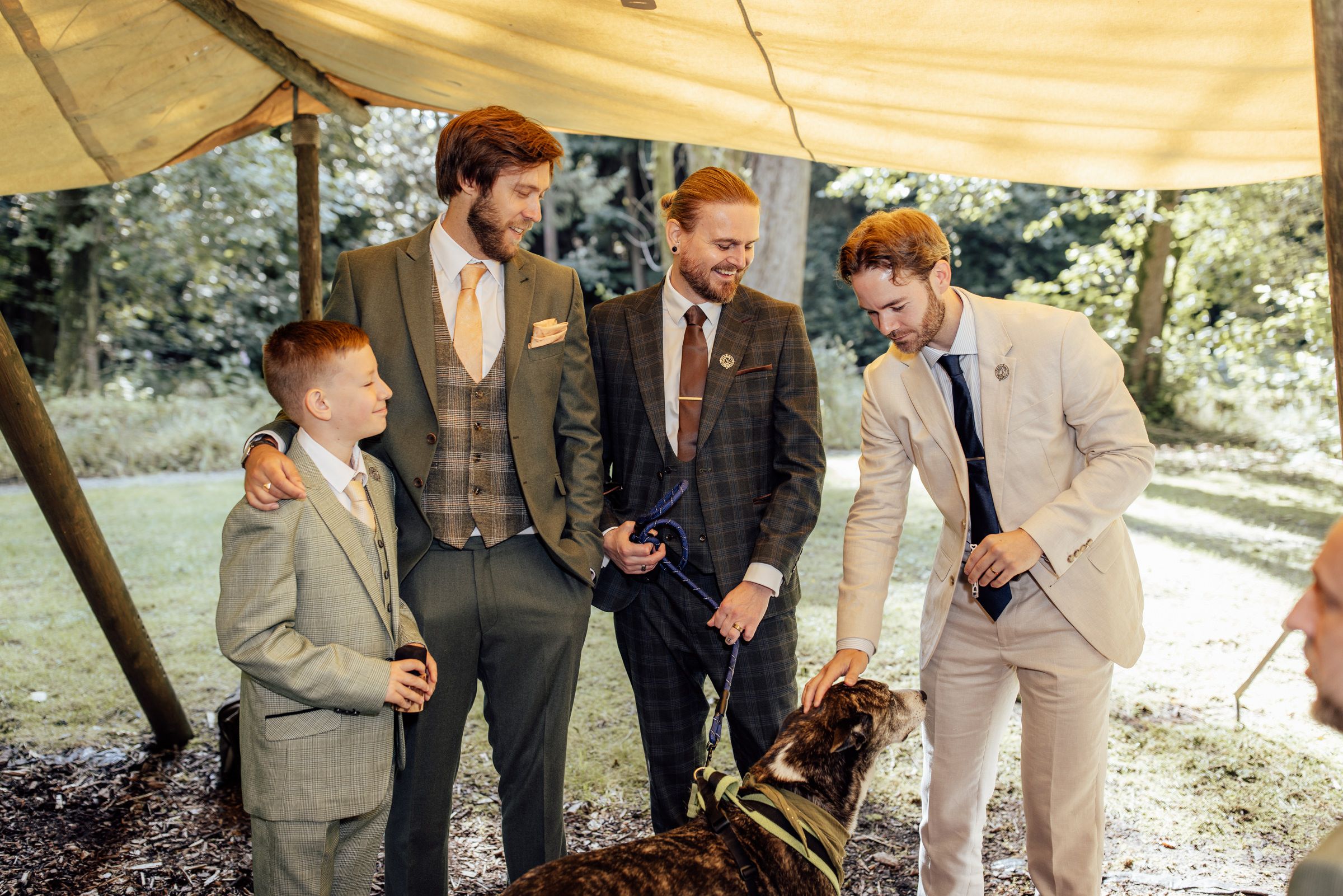 Groomsmen in tweed suits laughing with a child and bird of prey at outdoor wedding.