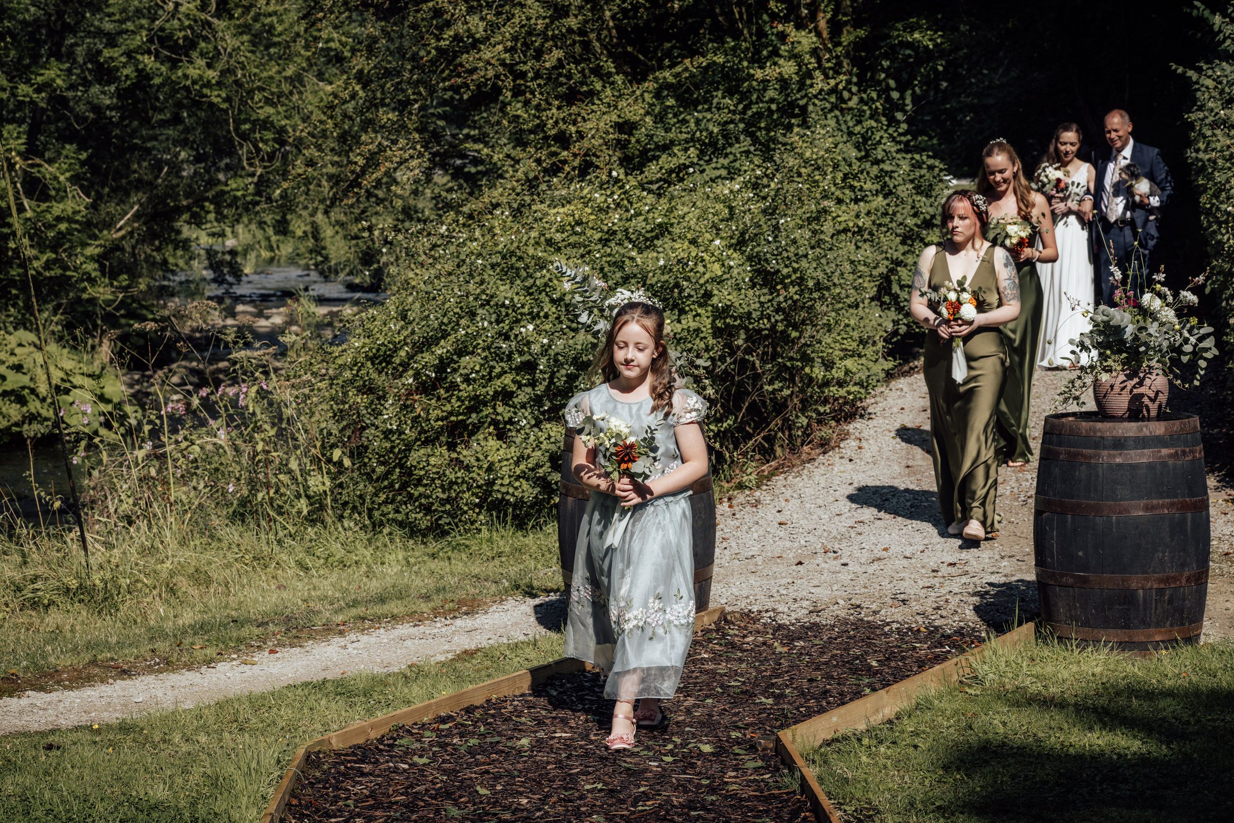 Young flower girl in blue dress walks down outdoor wedding aisle carrying bouquet, bridal party follows behind.