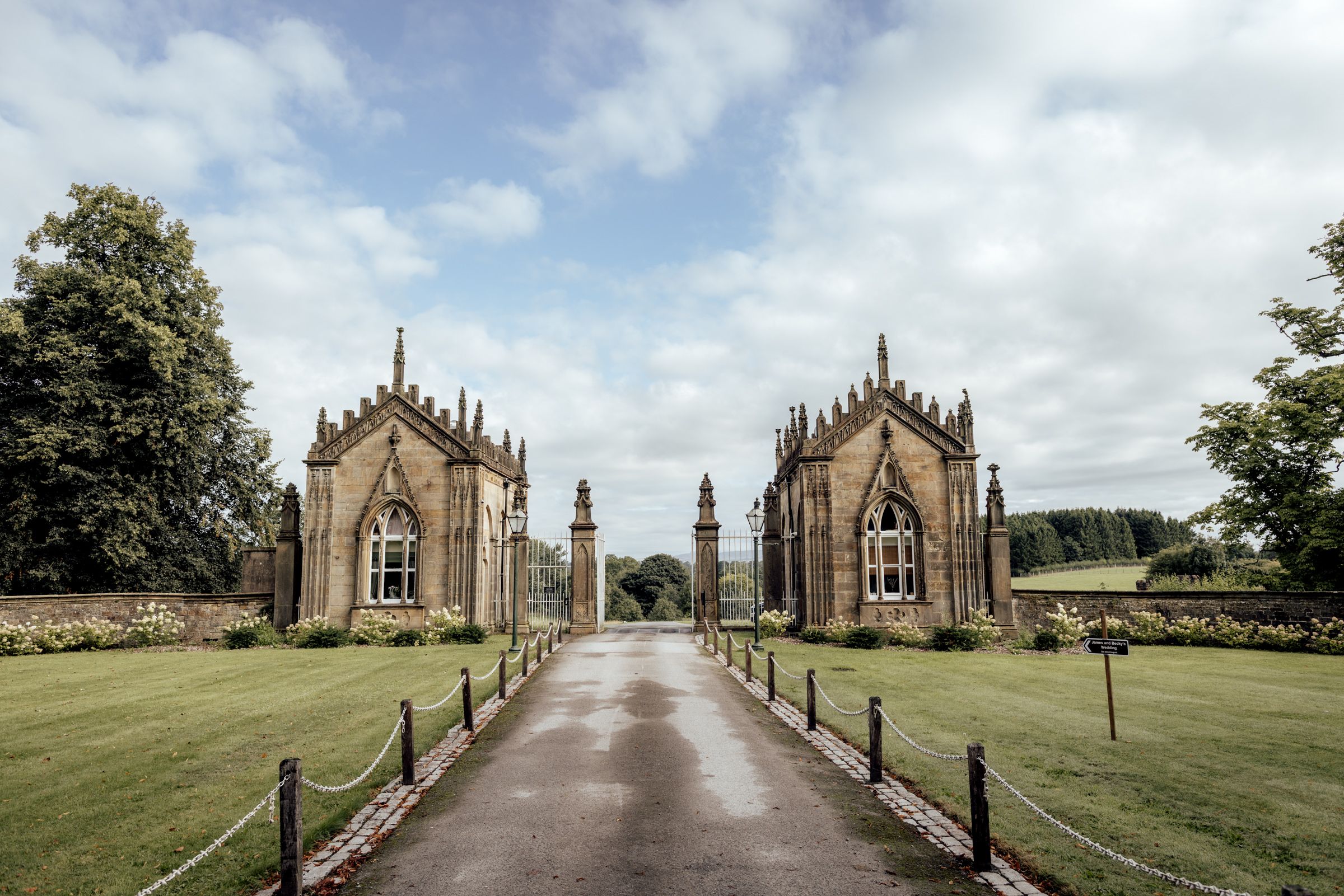 Gothic stone gate lodge entrance with a tree-lined driveway leading to a historic estate.