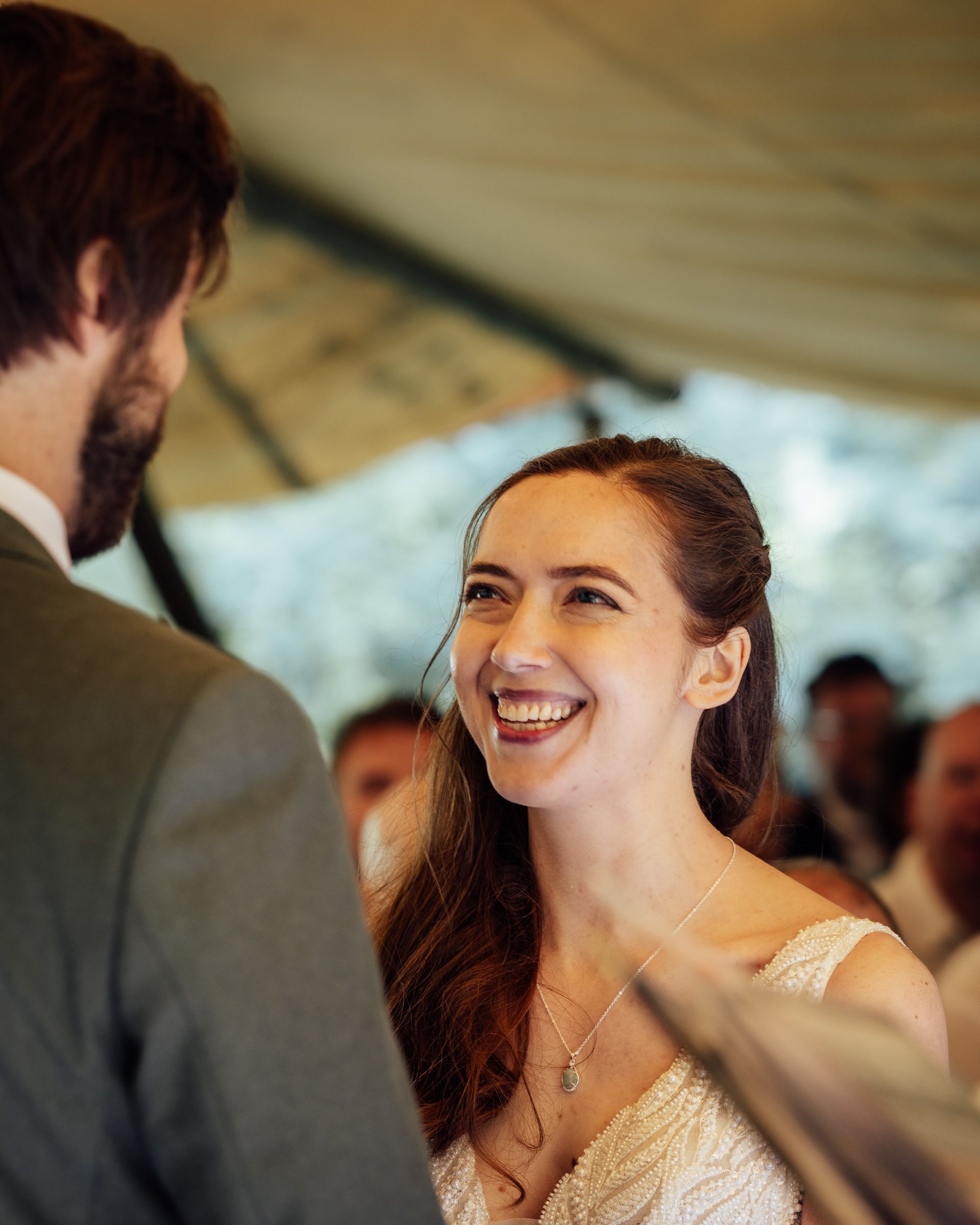 Bride smiling joyfully at groom during outdoor wedding ceremony under white tent.