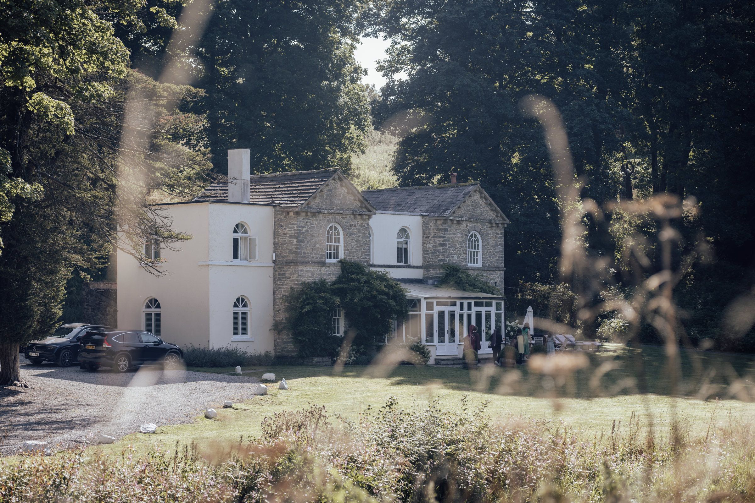 A grand country house surrounded by lush trees and manicured grounds on a sunny day.