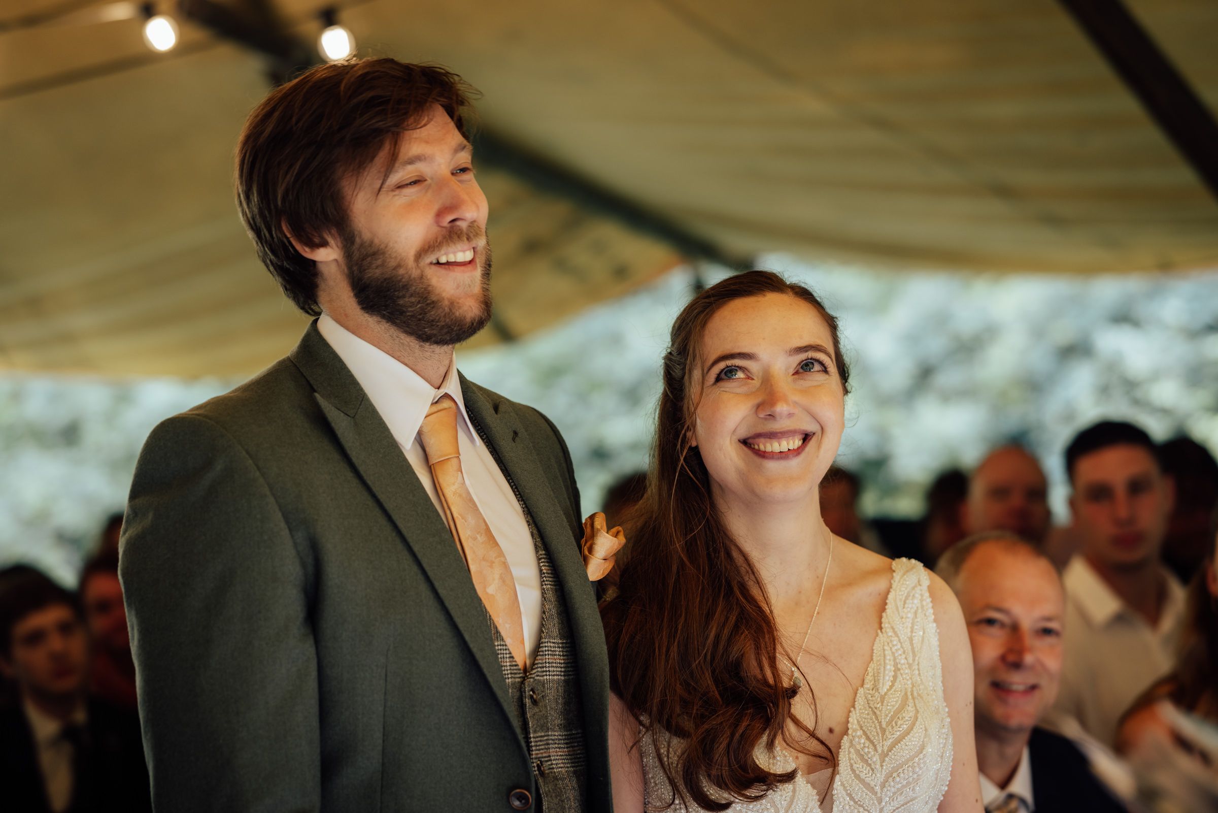 Happy couple smiling at wedding reception under a marquee tent, groom in gray suit, bride in white dress.