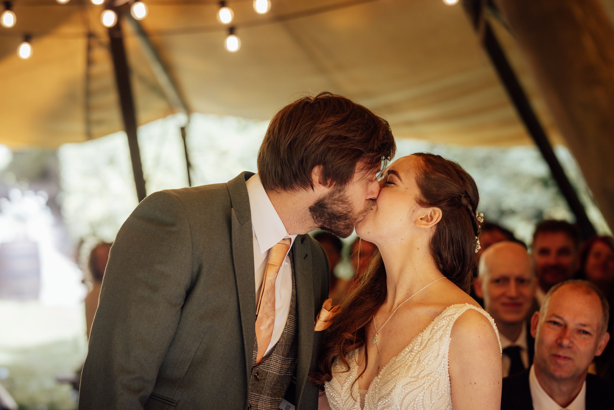 Bride and groom share first kiss at outdoor wedding ceremony under festoon lights.