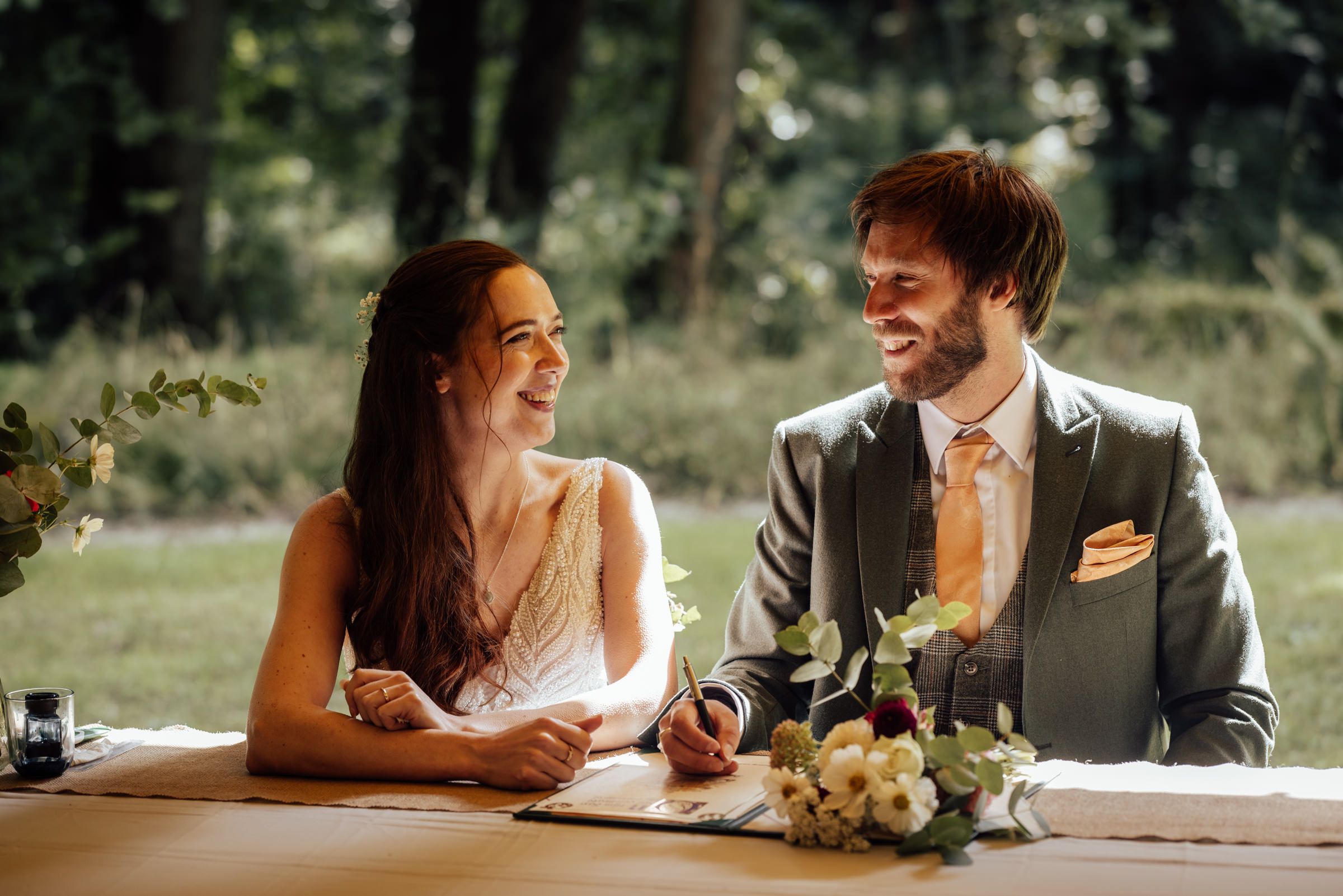 Happy couple signing wedding documents at an outdoor reception table with floral arrangements in a garden setting.