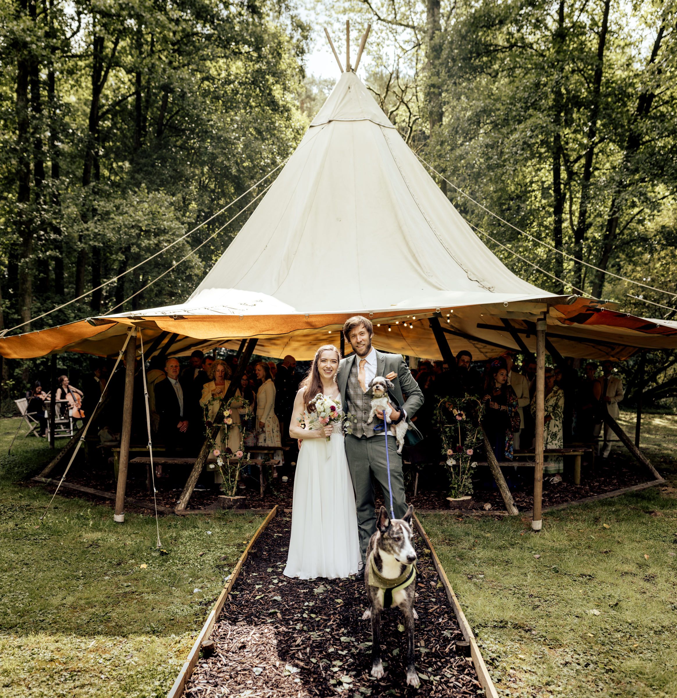 Bride and groom stand with their dog before a large tipi wedding tent surrounded by lush green trees.