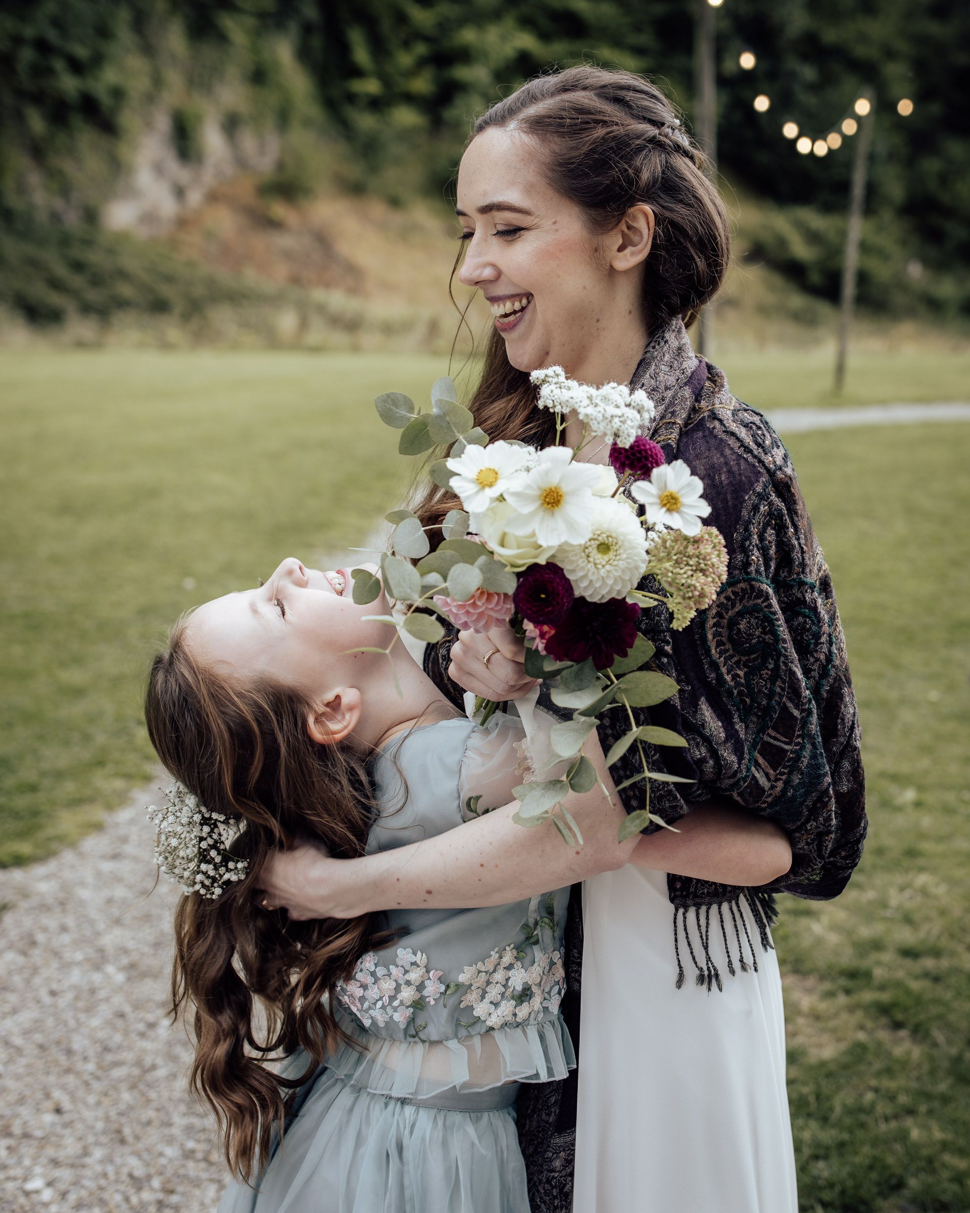 Bride laughing with flower girl holding white and burgundy wedding bouquet outdoors at garden wedding ceremony.
