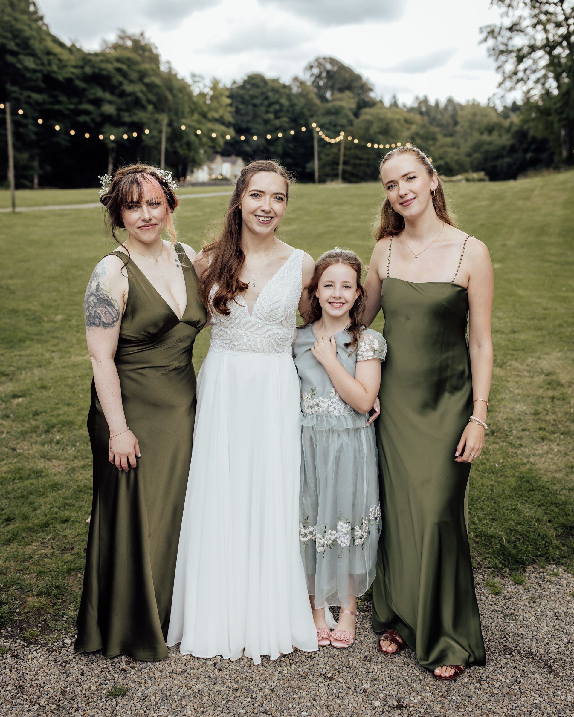 Bride in white gown poses with two bridesmaids in olive green dresses and a flower girl outdoors.
