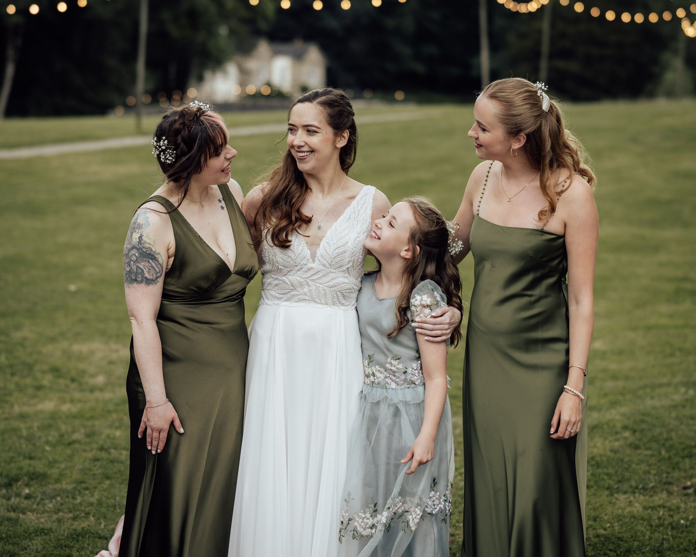 Bride in white gown with two bridesmaids in olive green dresses and flower girl laughing outdoors.