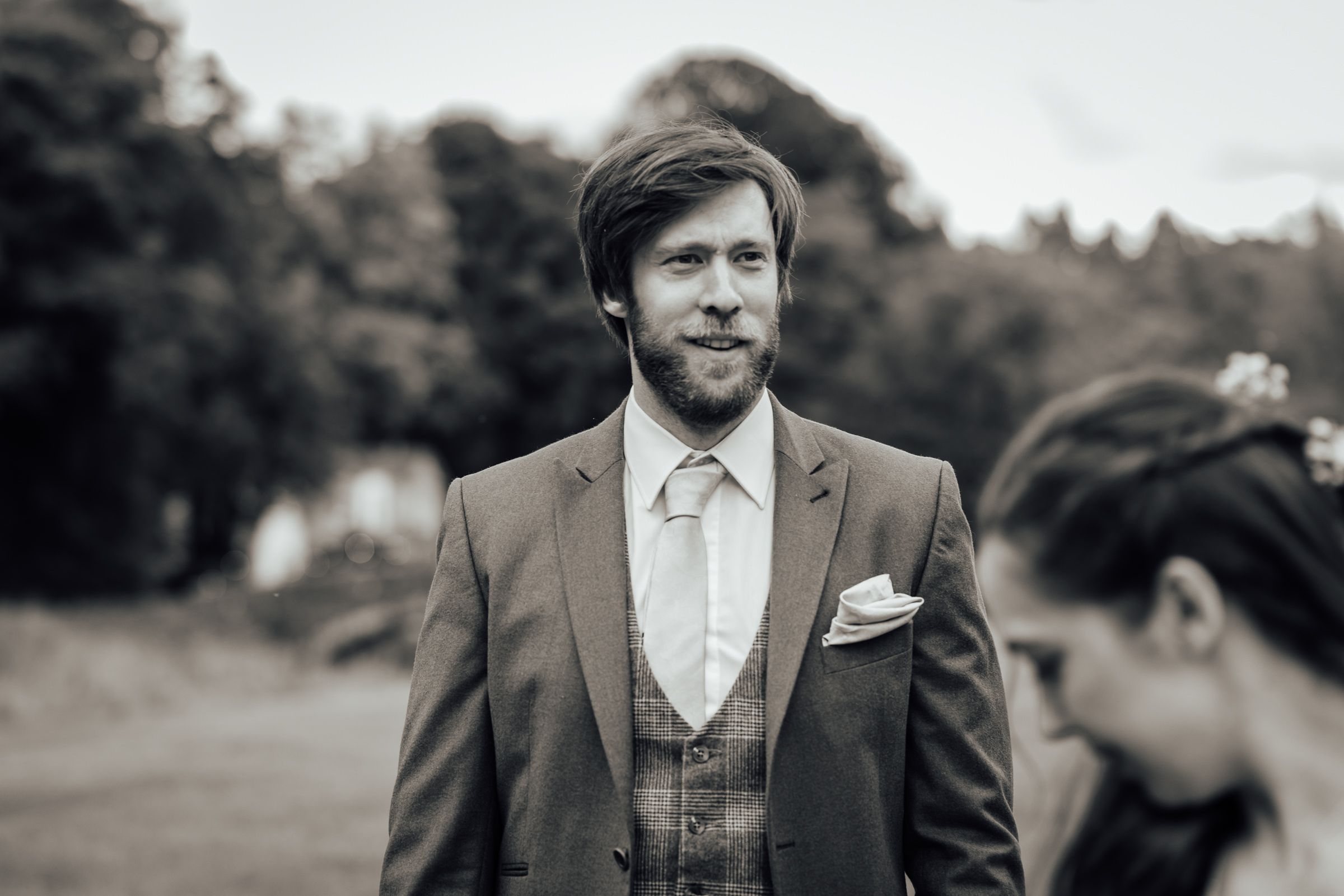 Smiling bearded groom in gray suit with white tie and pocket square at outdoor wedding ceremony.