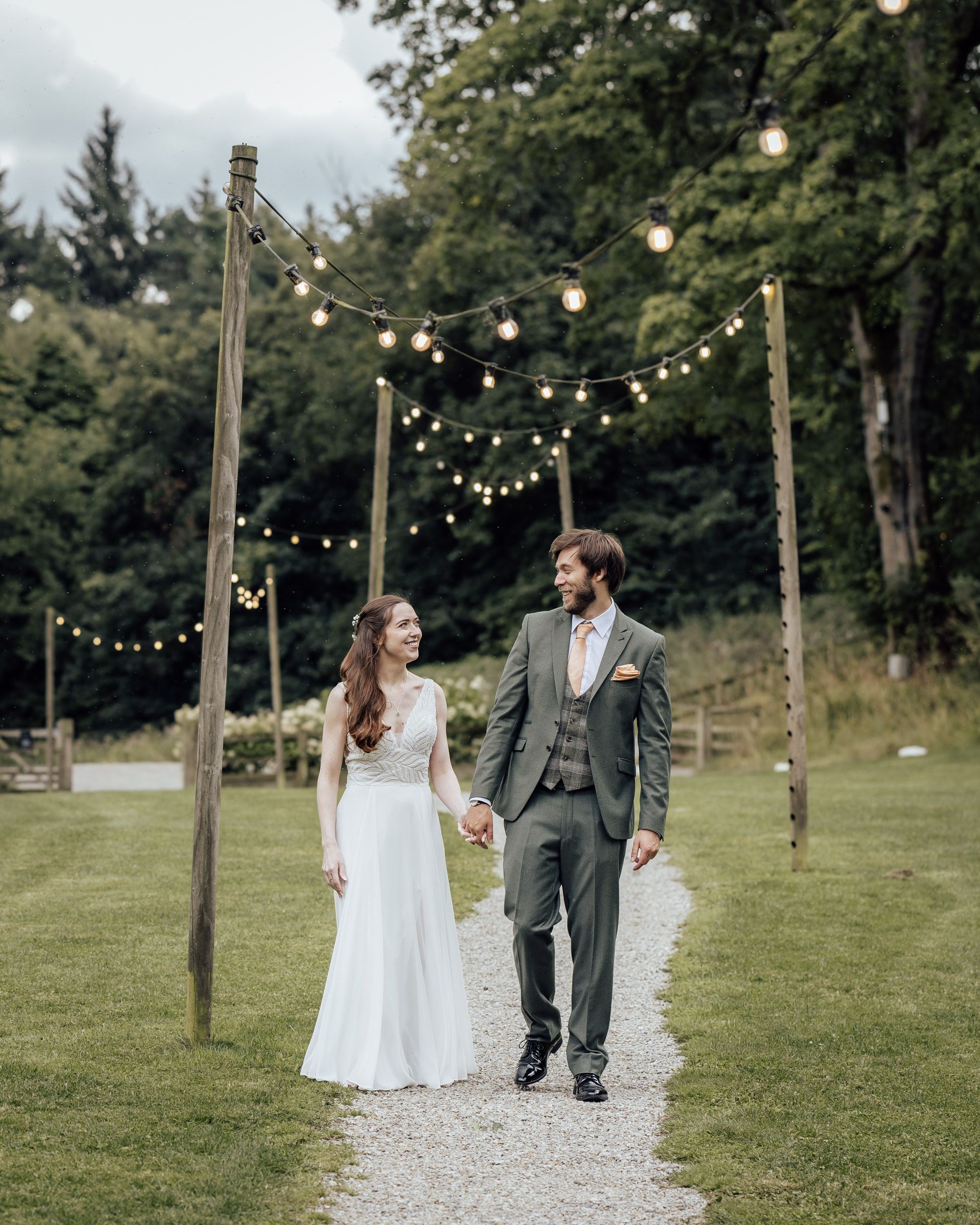 Bride and groom walking together under festoon lights strung between wooden posts at a lush green outdoor wedding venue.
