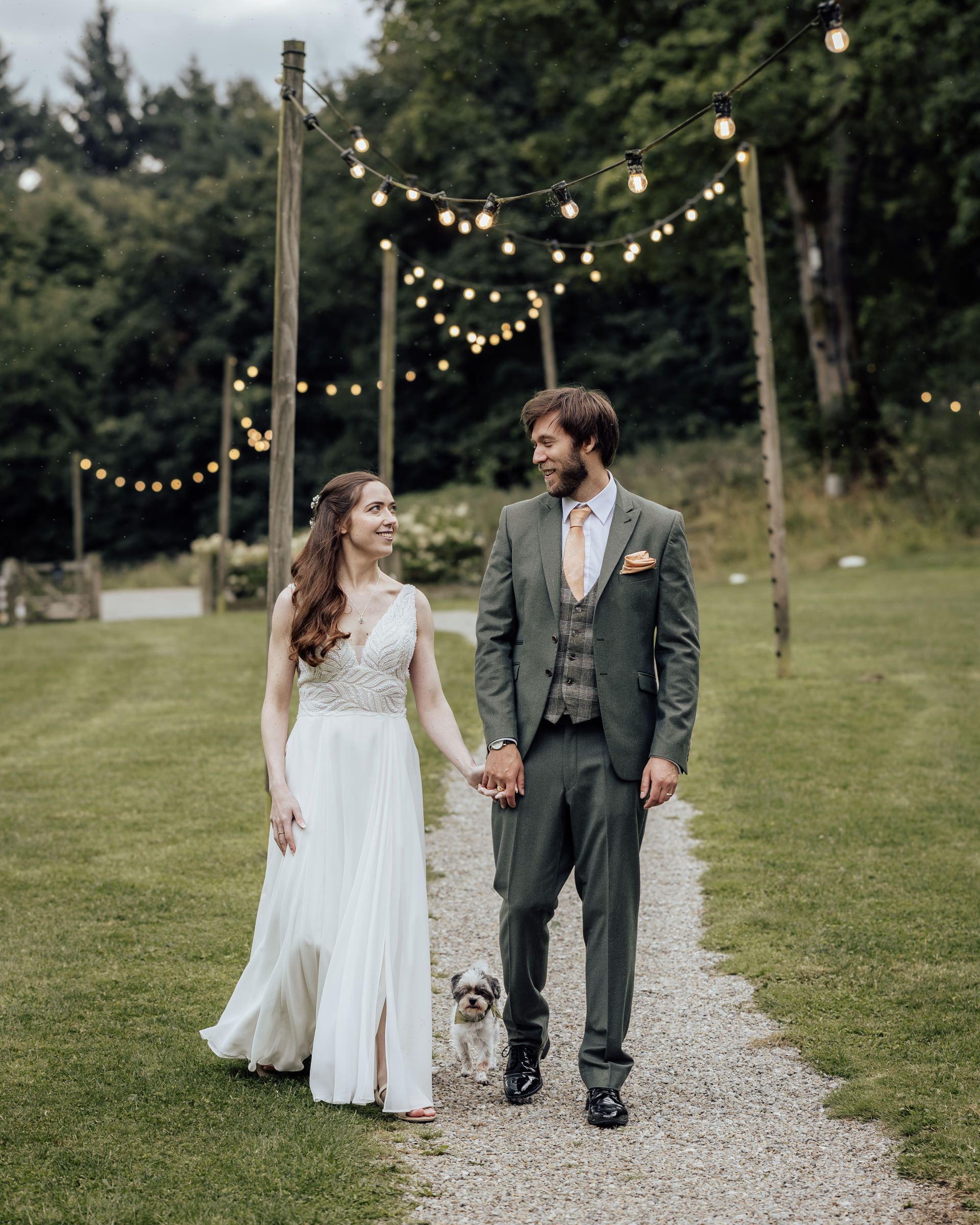 Bride and groom holding hands on a garden path with string lights and a small dog at their outdoor wedding.