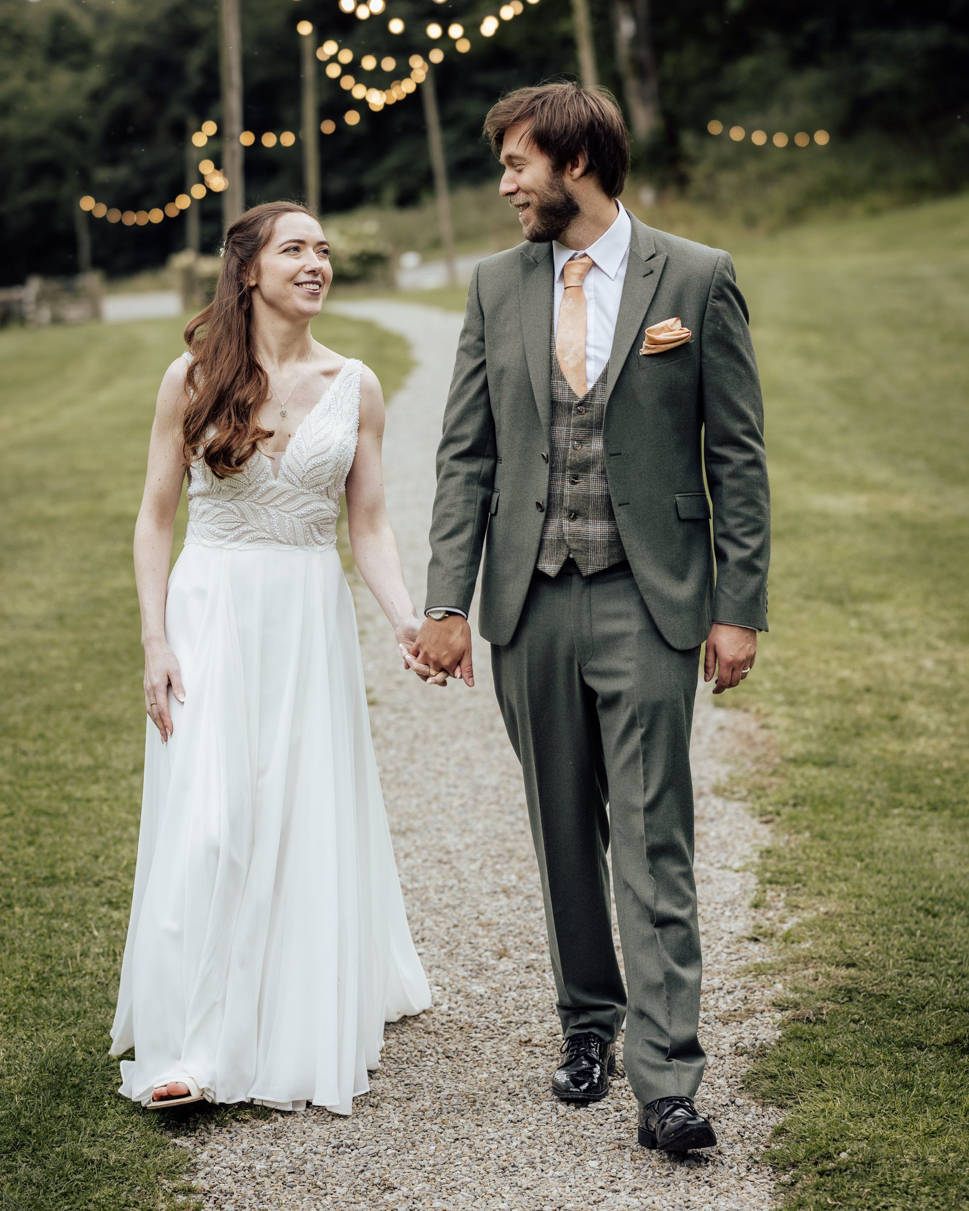 A bride and groom hold hands while walking on a garden path, with string lights and greenery in the background.
