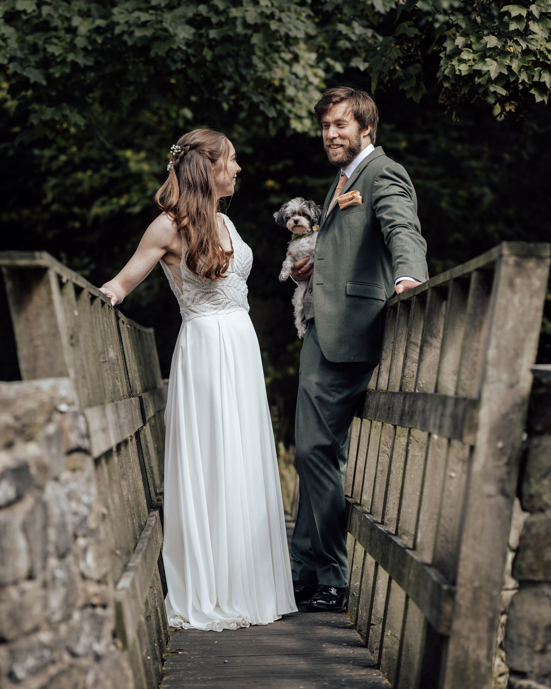 A bride and groom stand on a wooden bridge holding a small dog, surrounded by lush greenery on their wedding day.