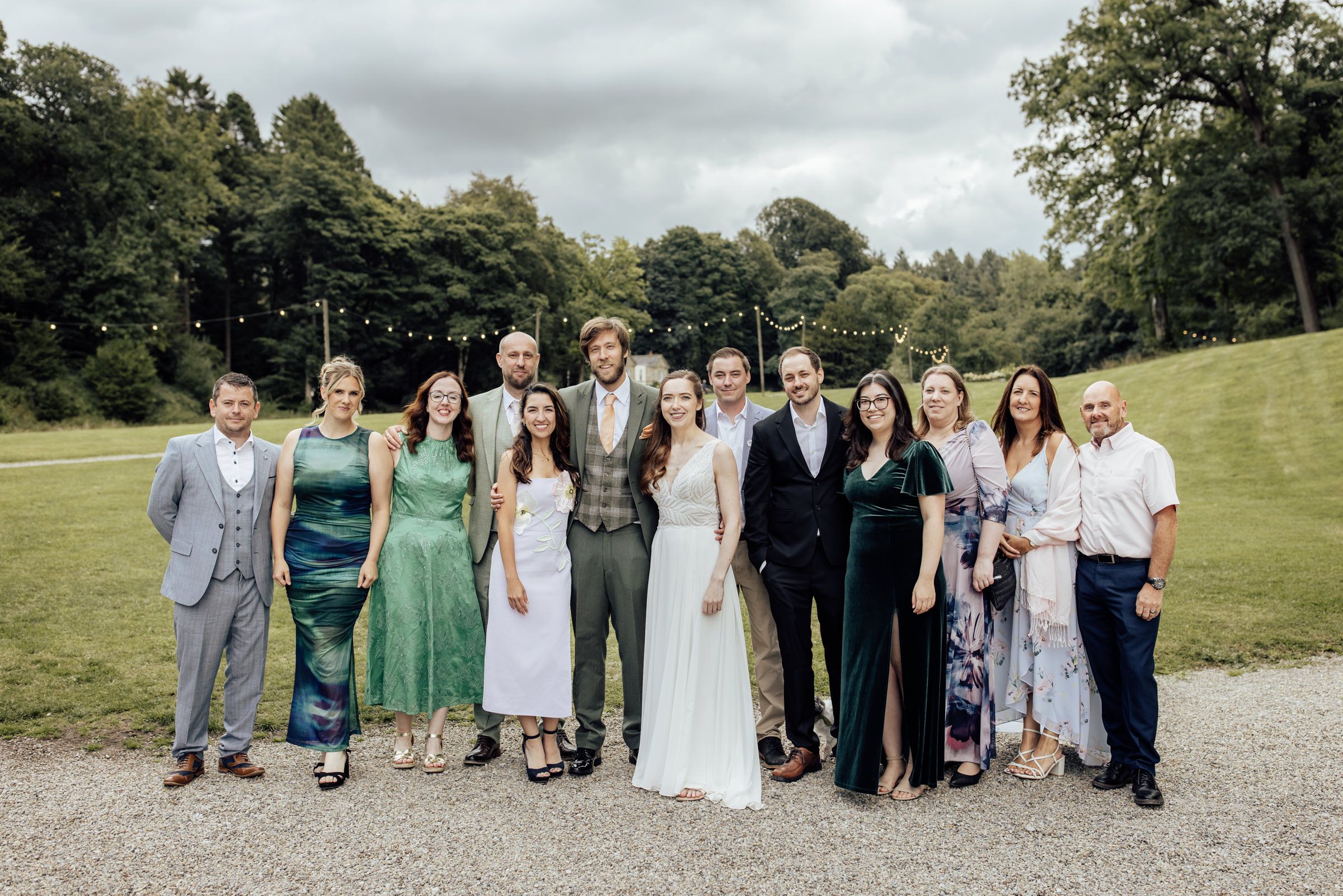Wedding party group photo outdoors on gravel path, guests in formal attire with bride in white gown, lush green trees backdrop.