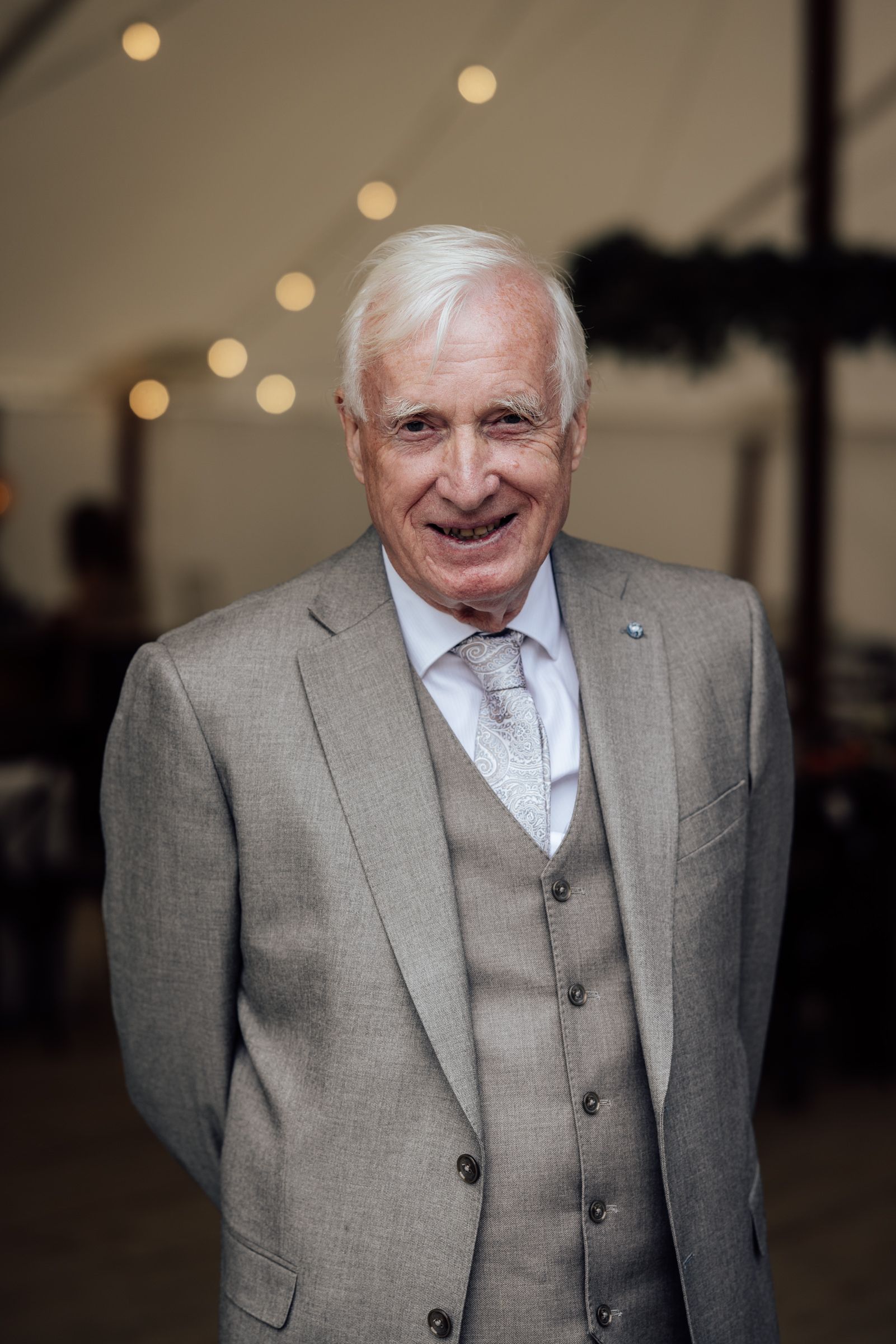 Elderly man in gray three-piece suit smiling at formal event with bokeh lights in background.