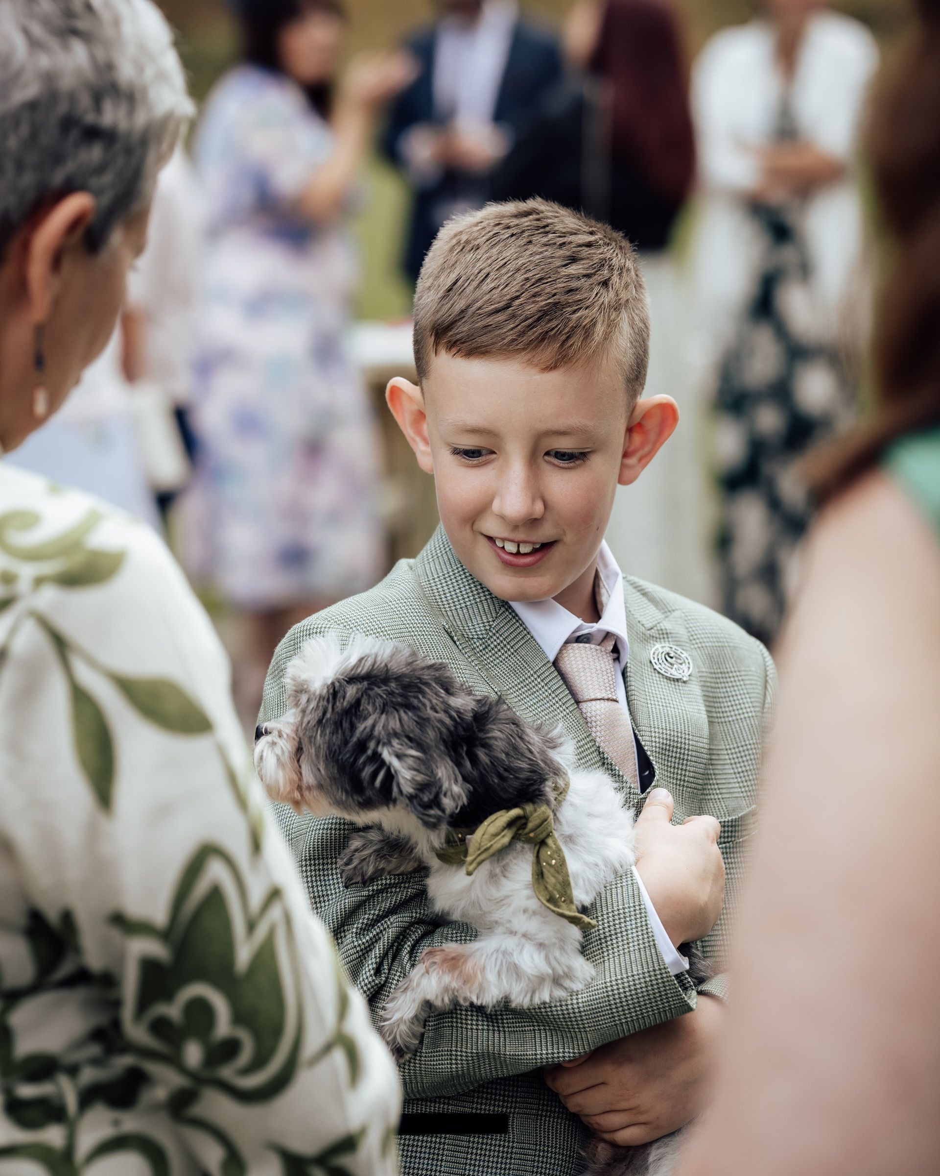 A smiling boy in a sage green suit holds a small fluffy puppy at an outdoor wedding celebration.