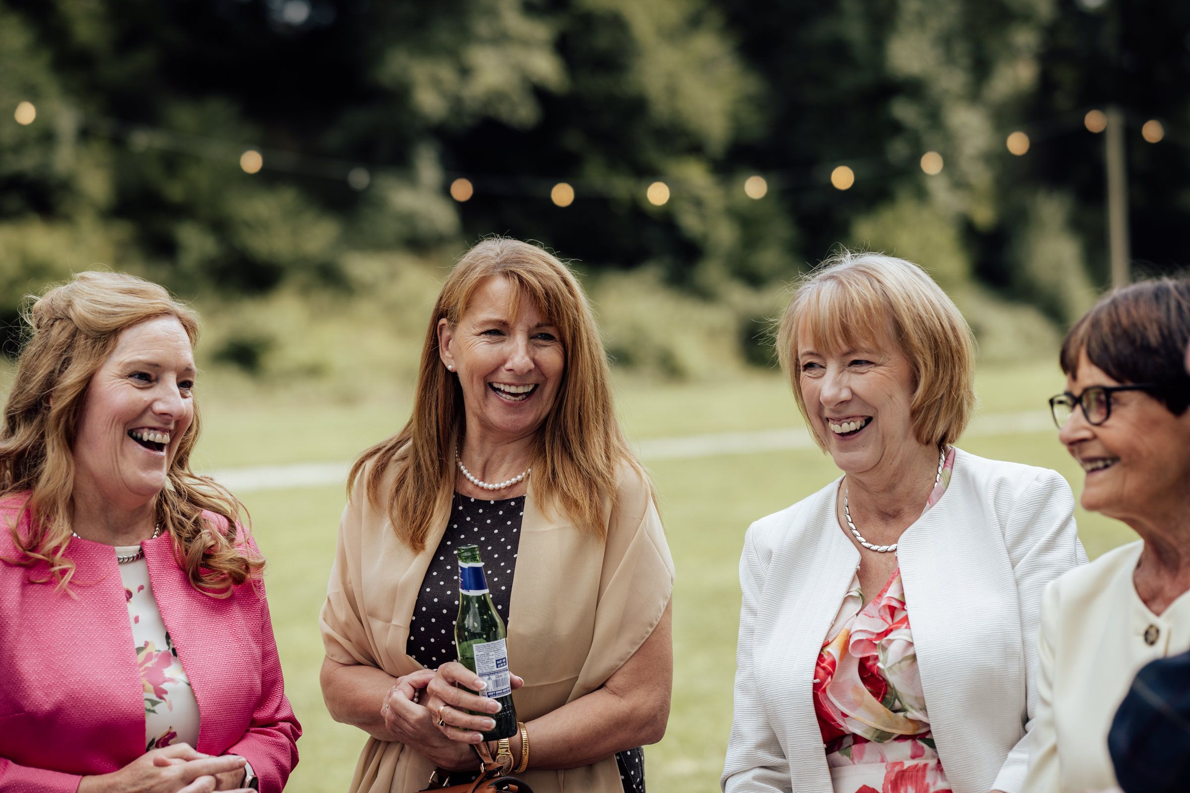 Four women laughing together outdoors at a garden party with string lights in the background.