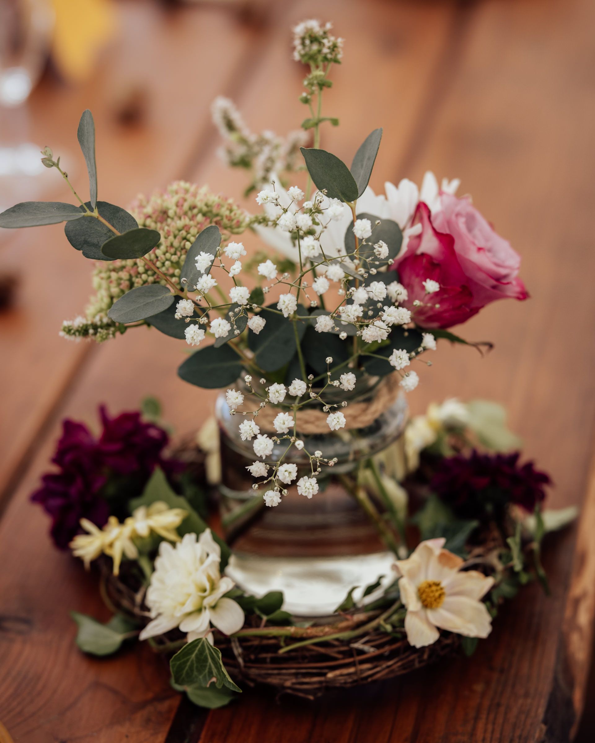 Rustic wedding centerpiece with pink roses, baby's breath, and eucalyptus in a mason jar on a twig wreath.