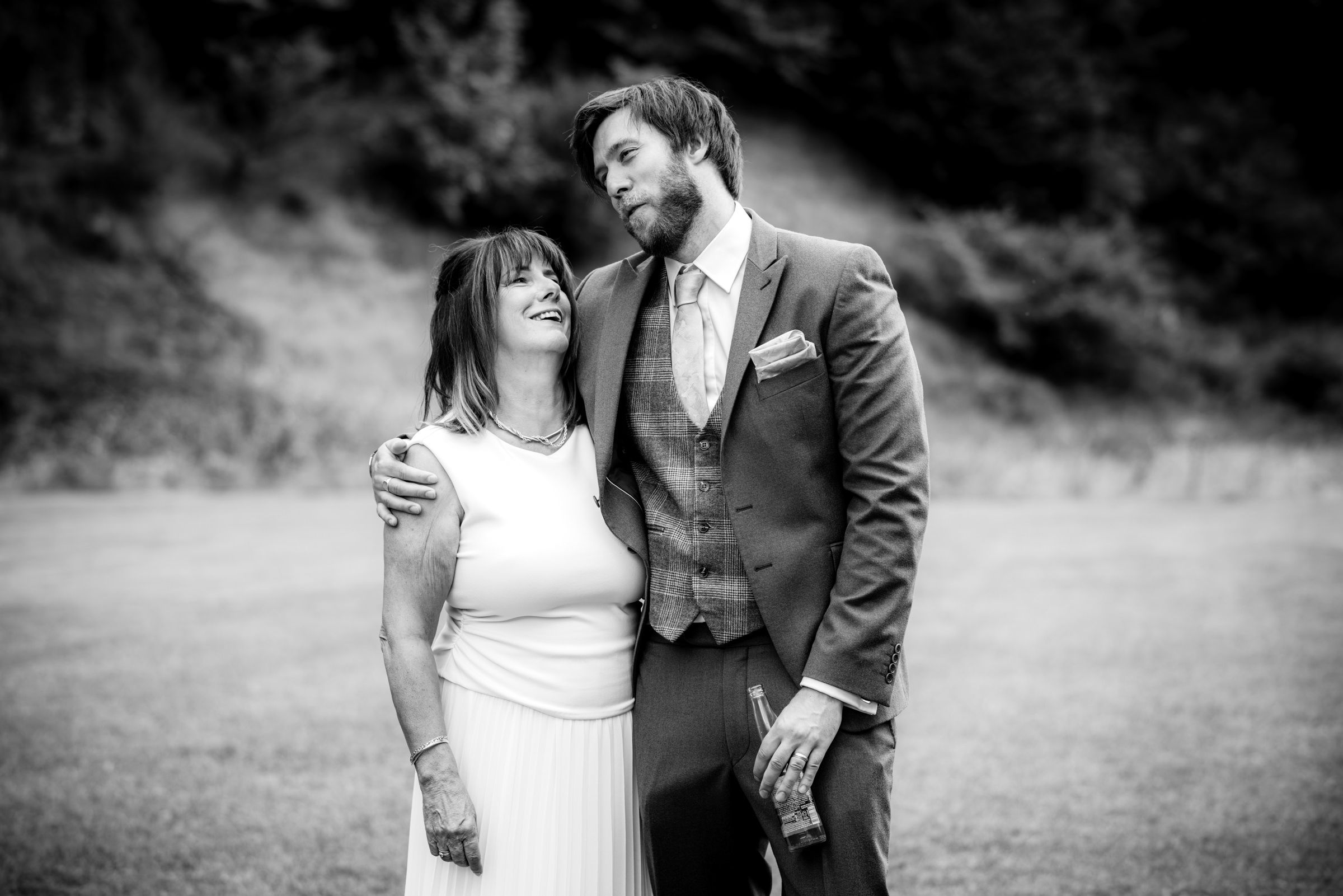 Black and white photo of a smiling couple at a wedding, man in suit with arm around woman in white dress.