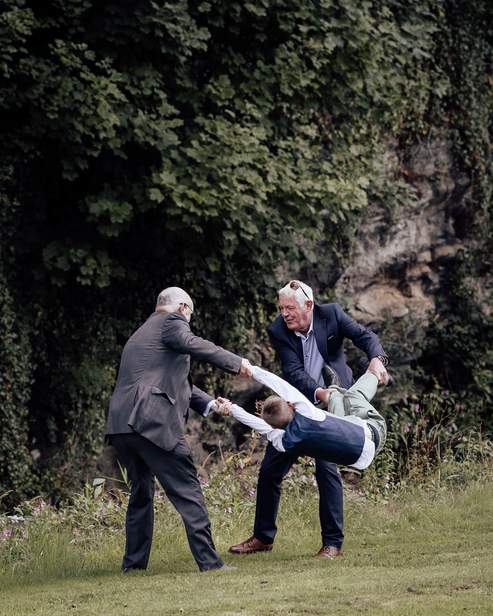 Two men playfully lift a boy in a suit outdoors at a wedding reception on a grass lawn.