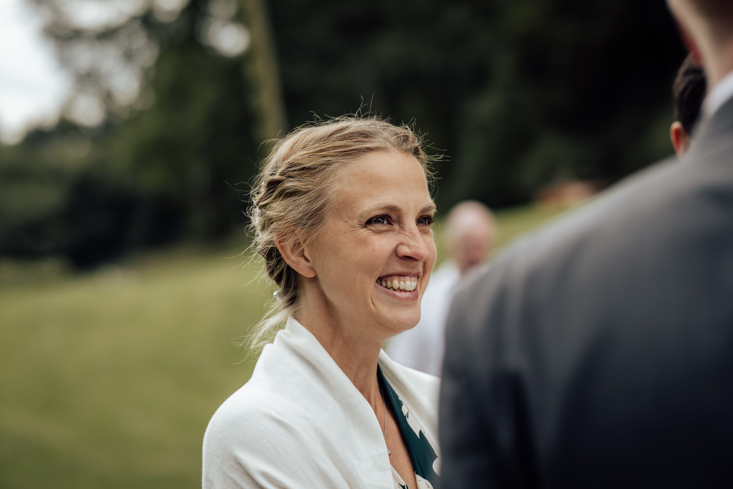 Smiling blonde woman in white jacket at outdoor wedding ceremony with green trees in background.
