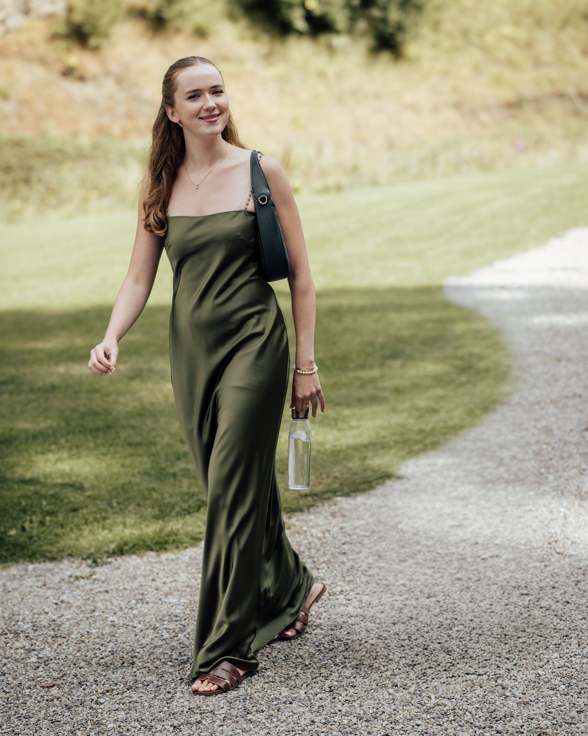 Woman in olive green satin maxi dress walking on gravel path, carrying clear bottle and green bag.