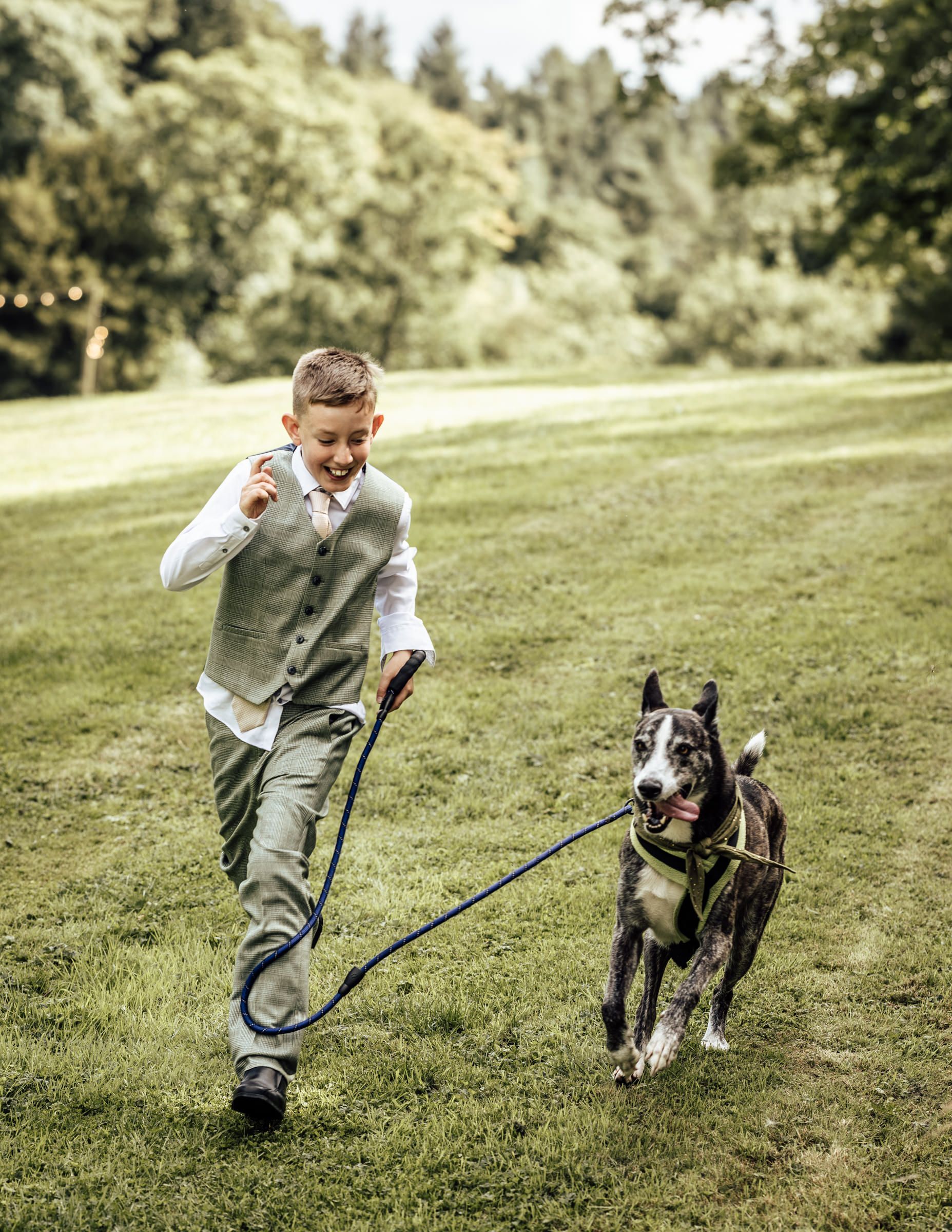 A joyful boy in a vest runs with a dog on a blue leash across a lush green lawn at an outdoor event.