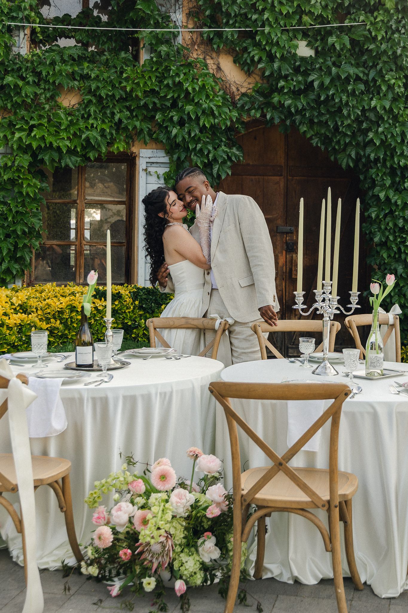 Interracial couple sharing a romantic kiss at an elegantly set outdoor wedding reception table adorned with candles and pink florals.