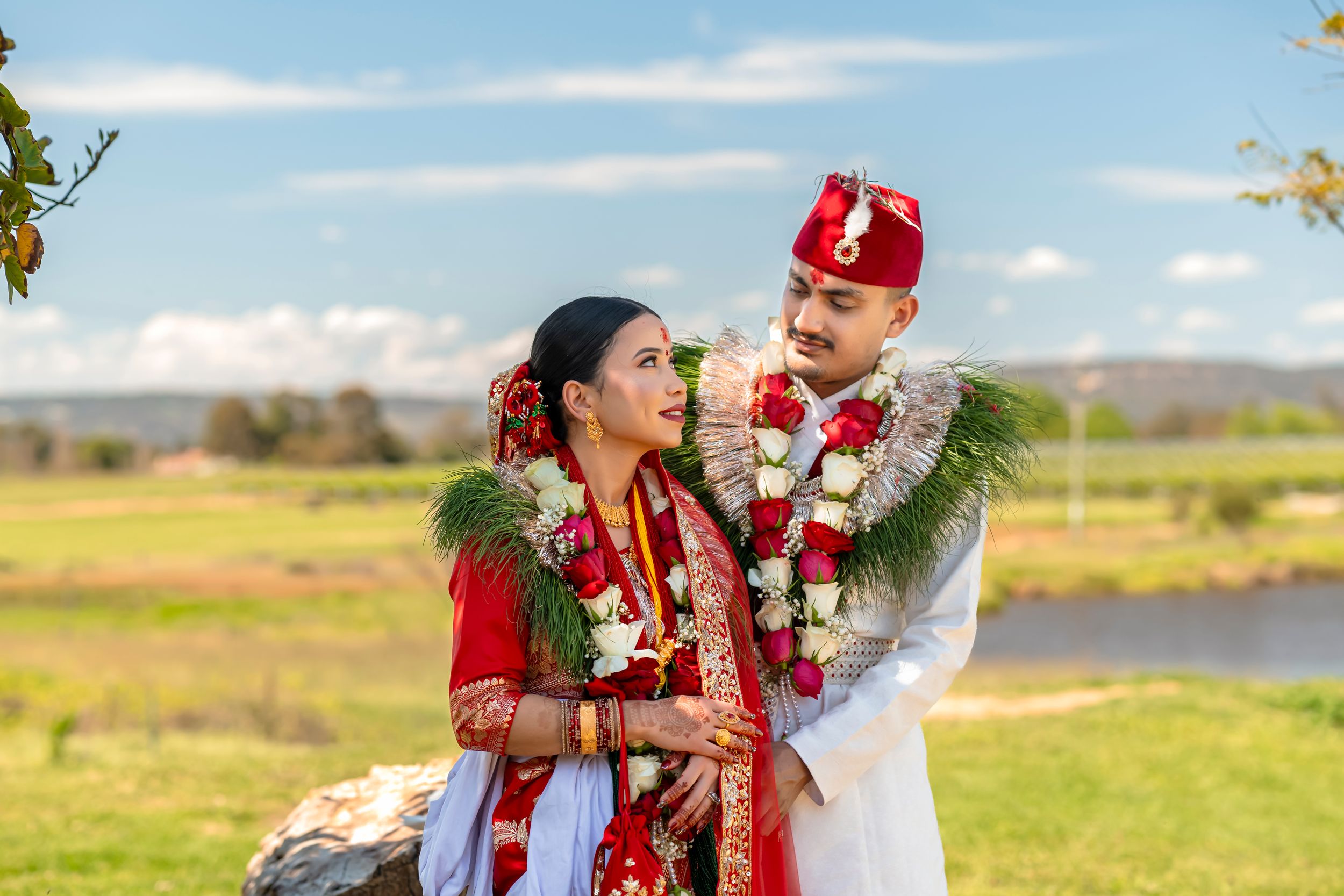 Couple in traditional Indian wedding attire with red and white garlands, posing outdoors under floral arch.