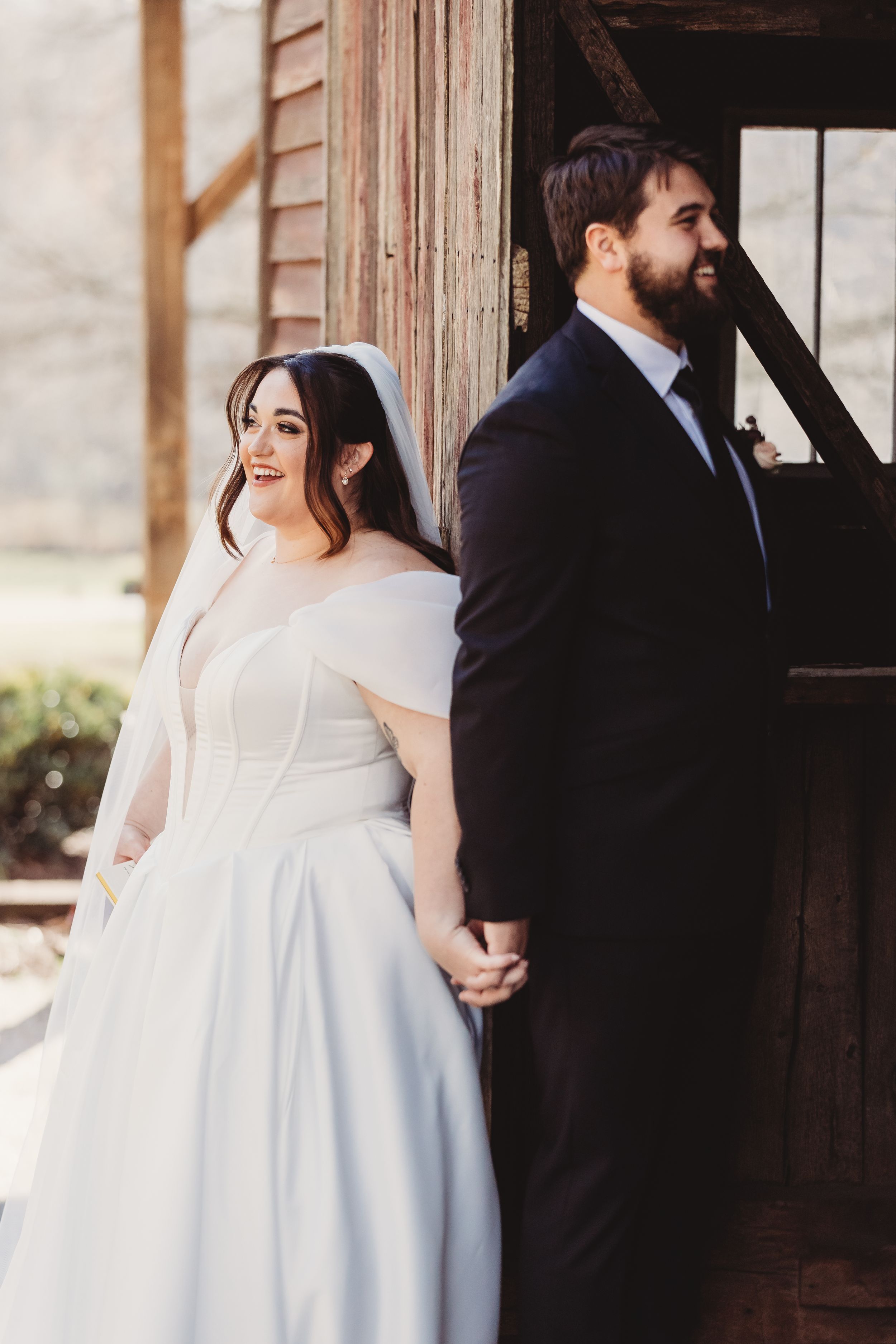 Bride in white off-shoulder gown and groom in black suit hold hands back-to-back at rustic barn wedding.