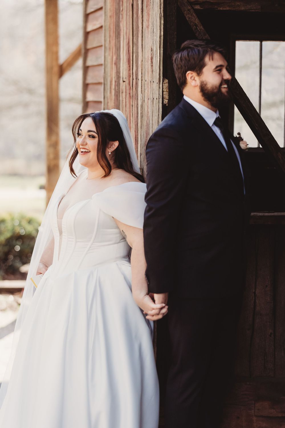 Bride in white off-shoulder gown and groom in black suit hold hands back-to-back at rustic barn wedding.