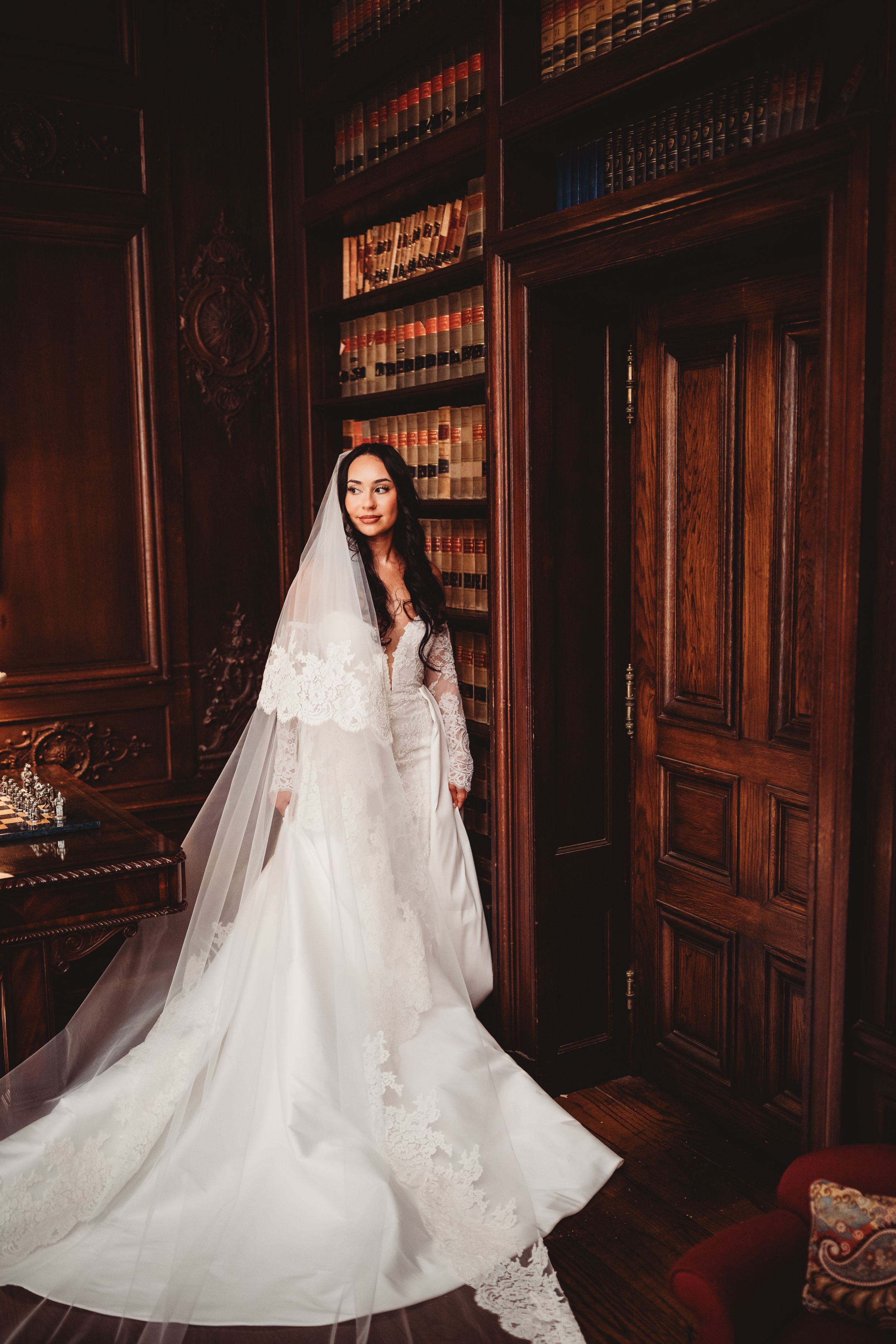 Bride in elegant lace wedding gown with veil poses in a dark wood-paneled library with bookshelves.