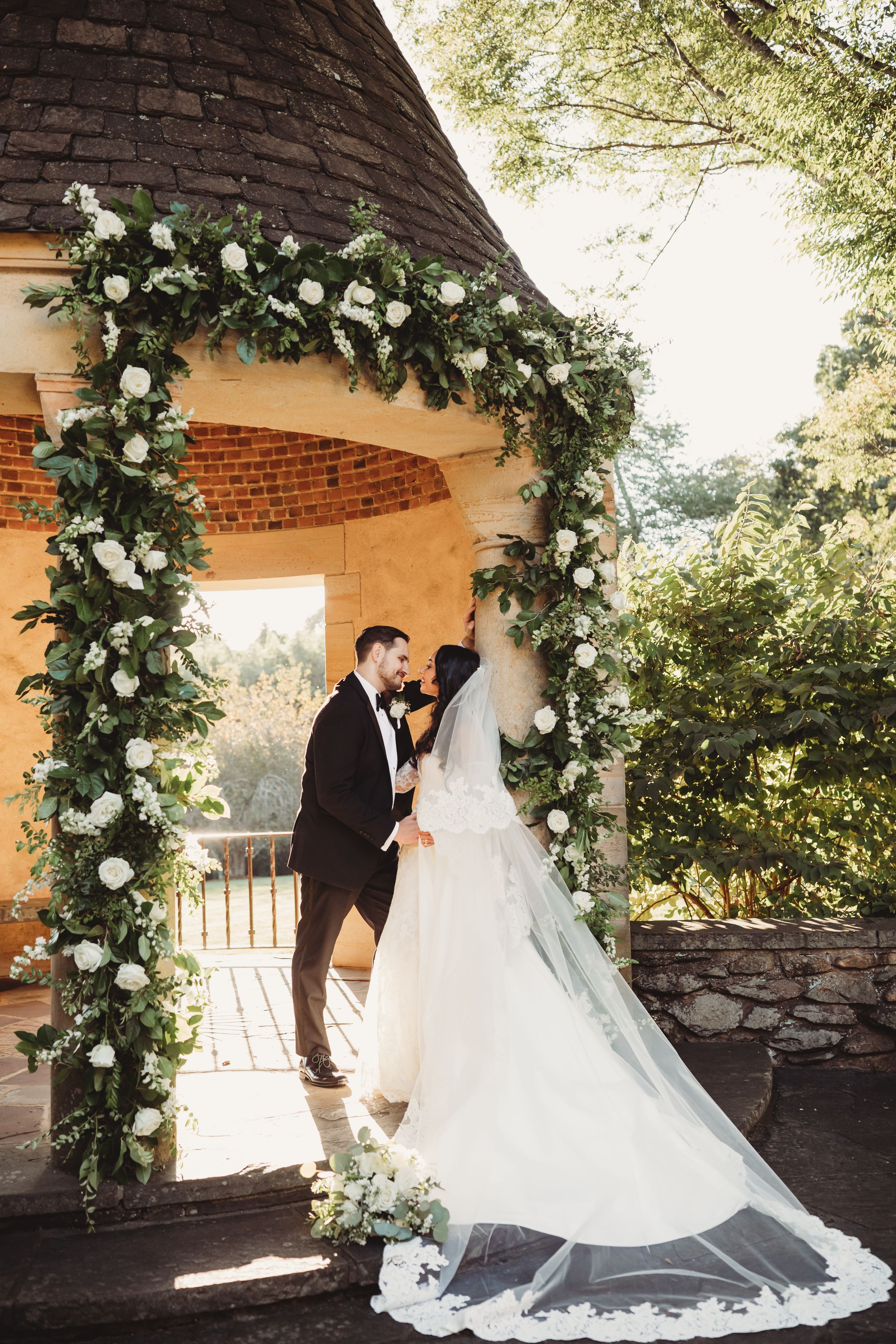 Bride and groom share a romantic moment under a floral arch adorned with white roses and greenery at an outdoor wedding.