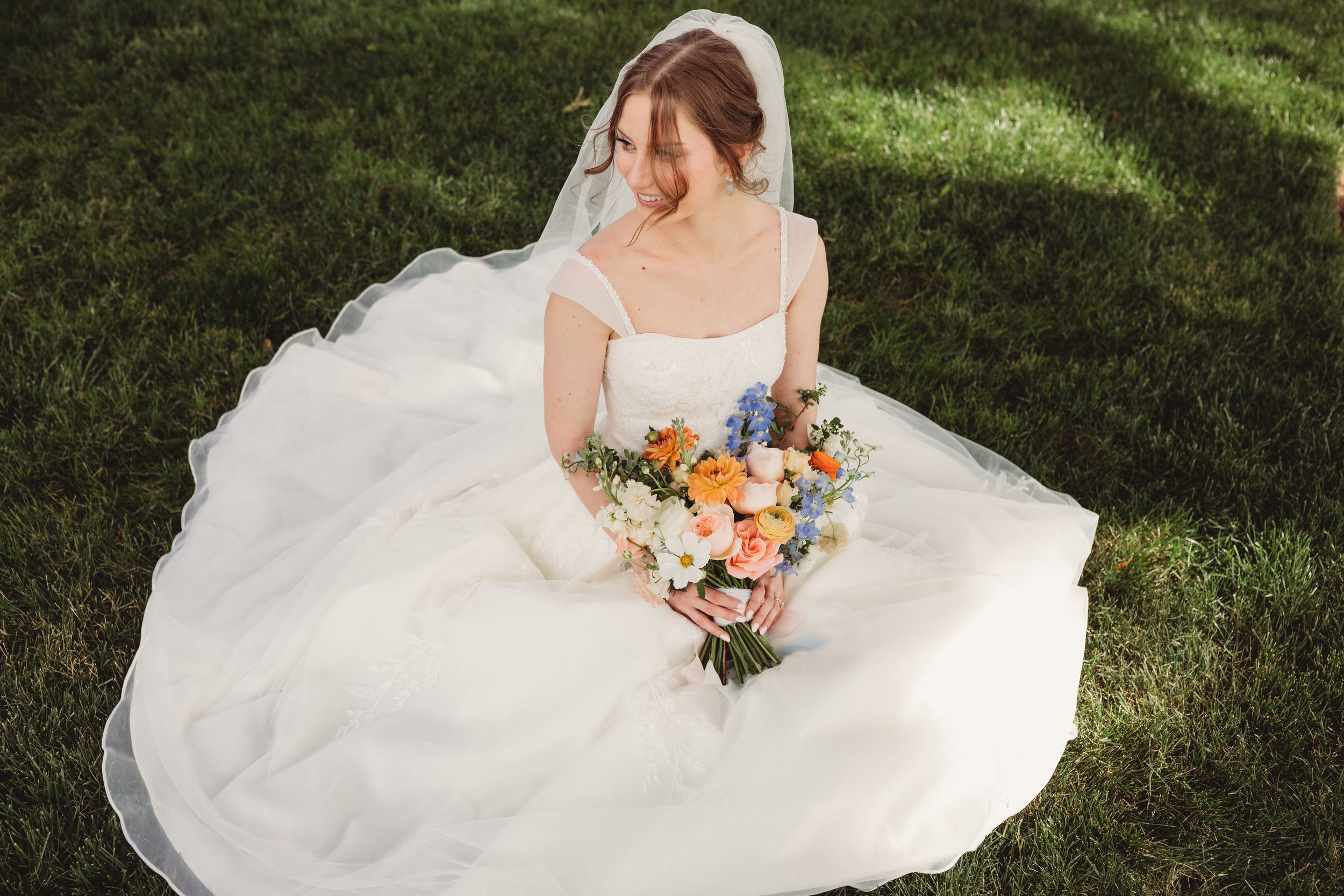 Bride in white ball gown and veil sits on green grass holding colorful bouquet of orange, pink, and blue flowers.
