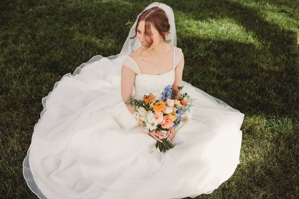 Bride in white ball gown and veil sits on green grass holding colorful bouquet of orange, pink, and blue flowers.