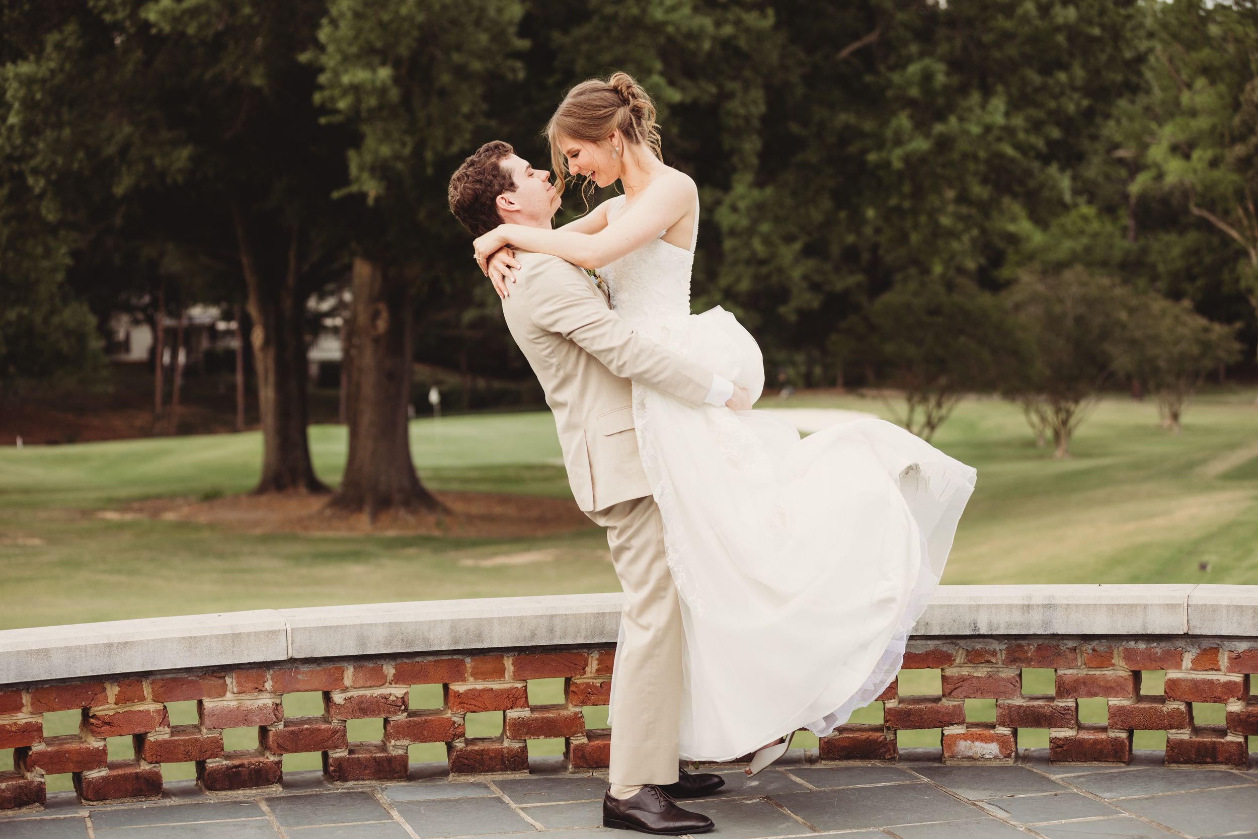Groom lifting bride in white wedding dress outdoors near brick wall with green trees in background.